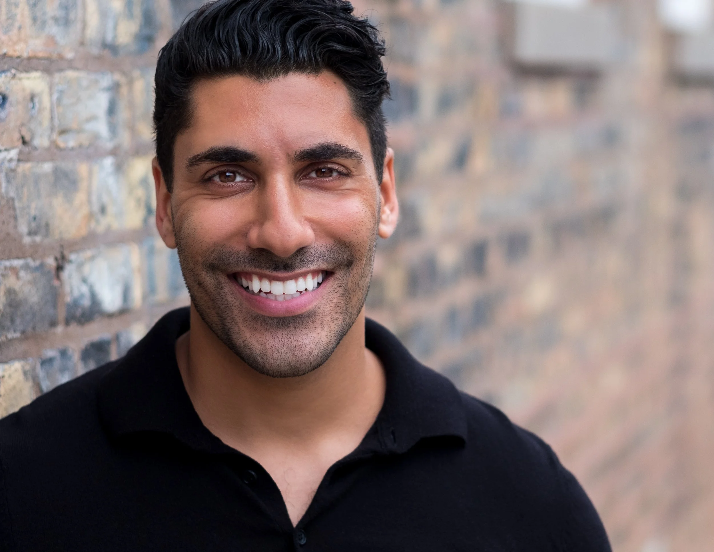 Professional headshot by St. Paul photographer Jay Cupcake. A smiling man with dark, wavy hair, a beard, wearing a black shirt, standing next to a brick wall.