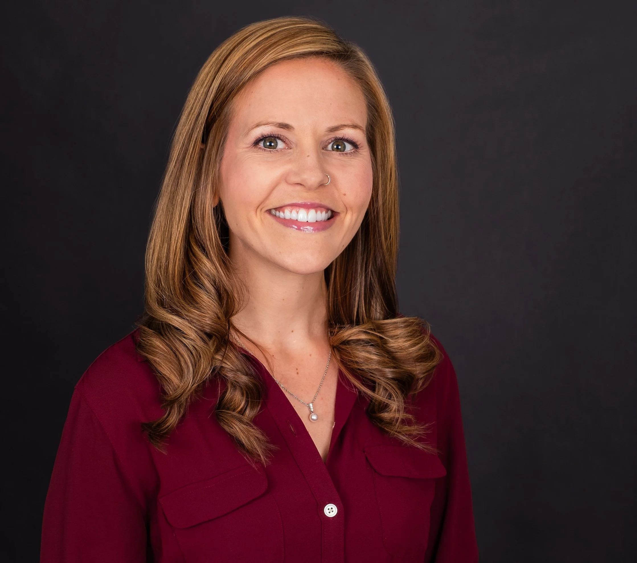 Professional Corporate Headshot by Minneapolis Photographer Jay Cupcake. A woman with long, wavy auburn hair and a light skin tone, smiling, wearing a maroon blouse and a silver necklace, against a dark background.