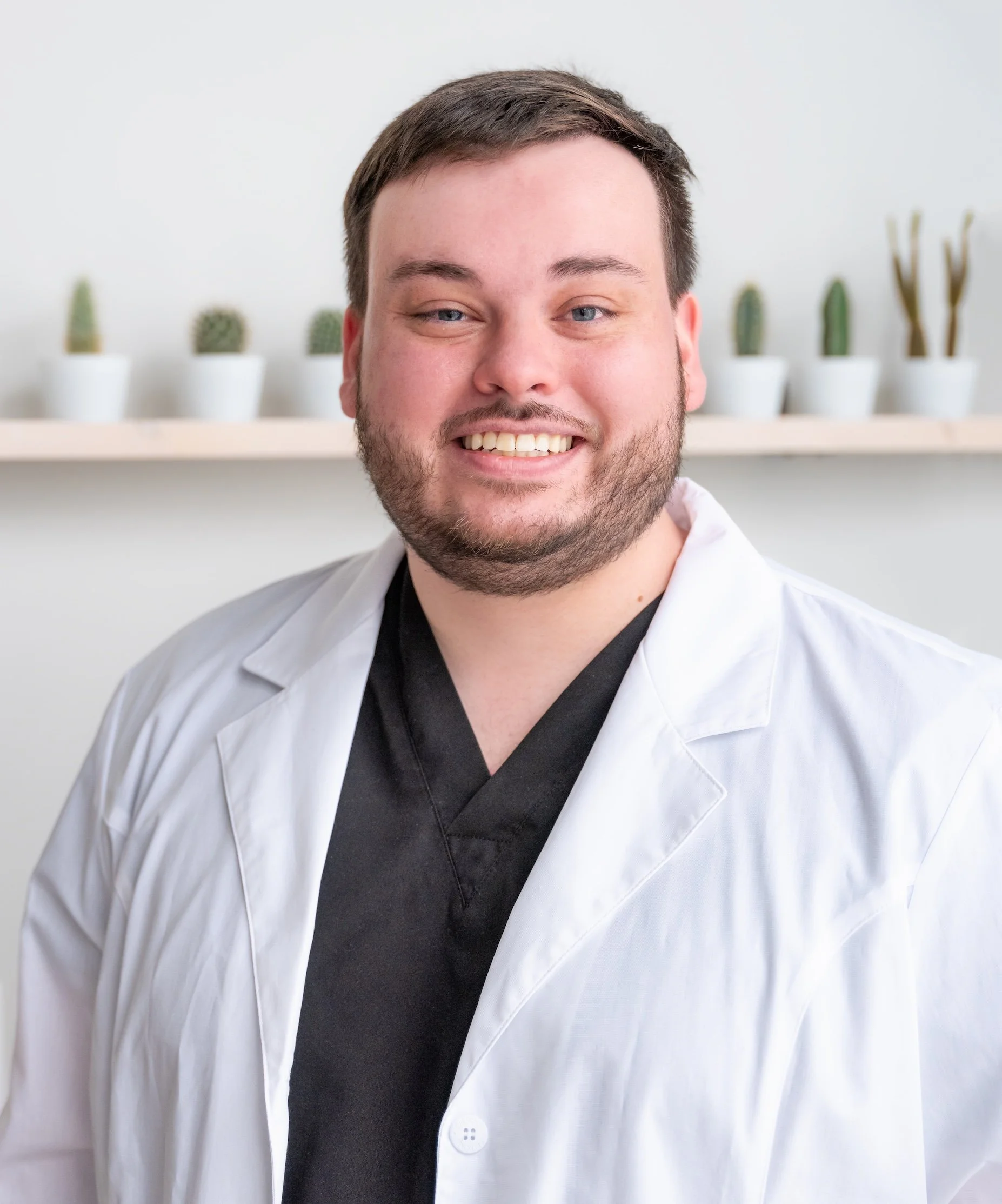 Professional Corporate Headshot by Saint Paul Photographer Jay Cupcake. A smiling man dressed in a white lab coat and black scrubs standing in front of a shelf with potted cacti.