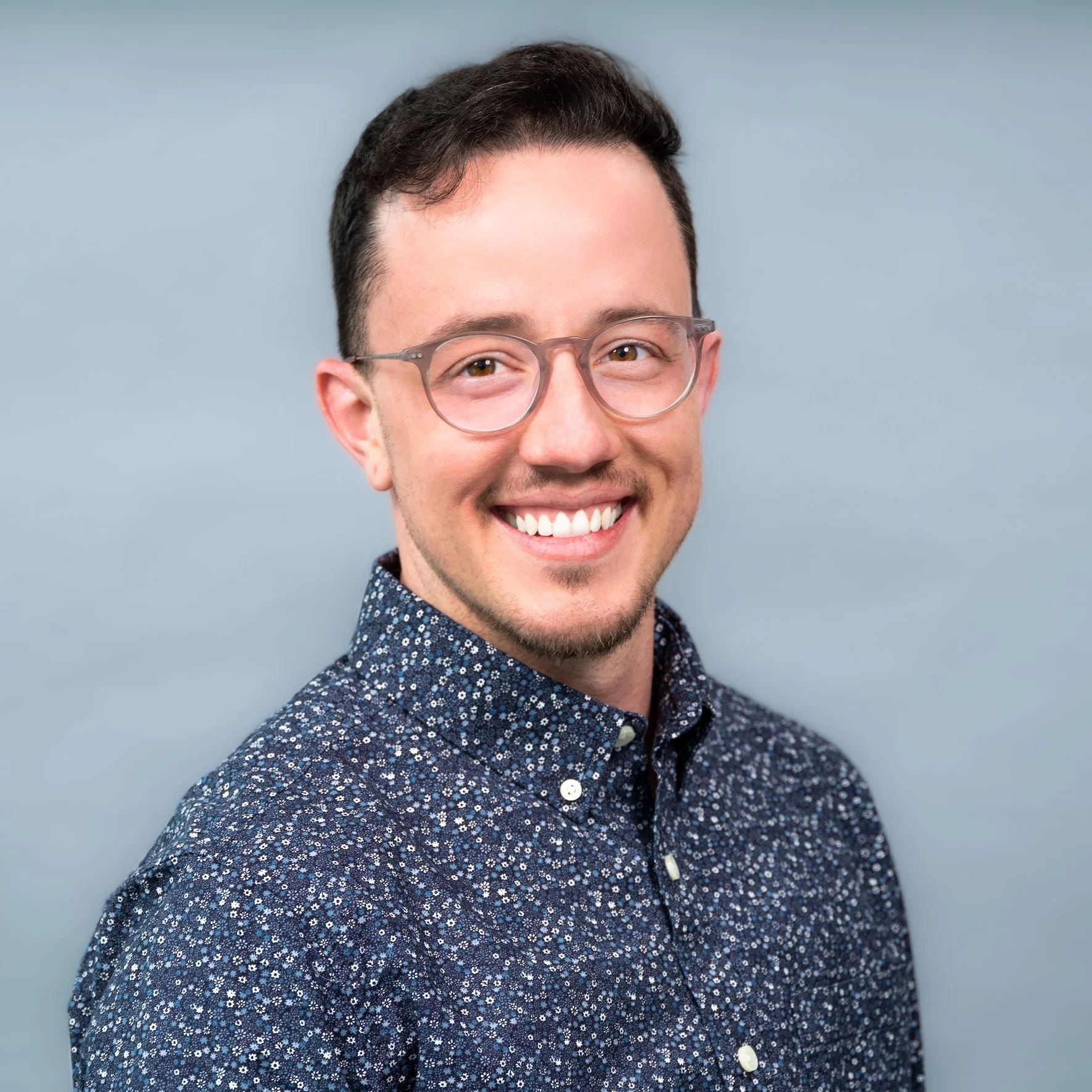 Professional Corporate Headshot by Saint Paul Photographer Jay Cupcake. A smiling young man with short dark hair, wearing clear glasses and a navy blue button-up shirt with small white floral patterns, standing against a light blue background.