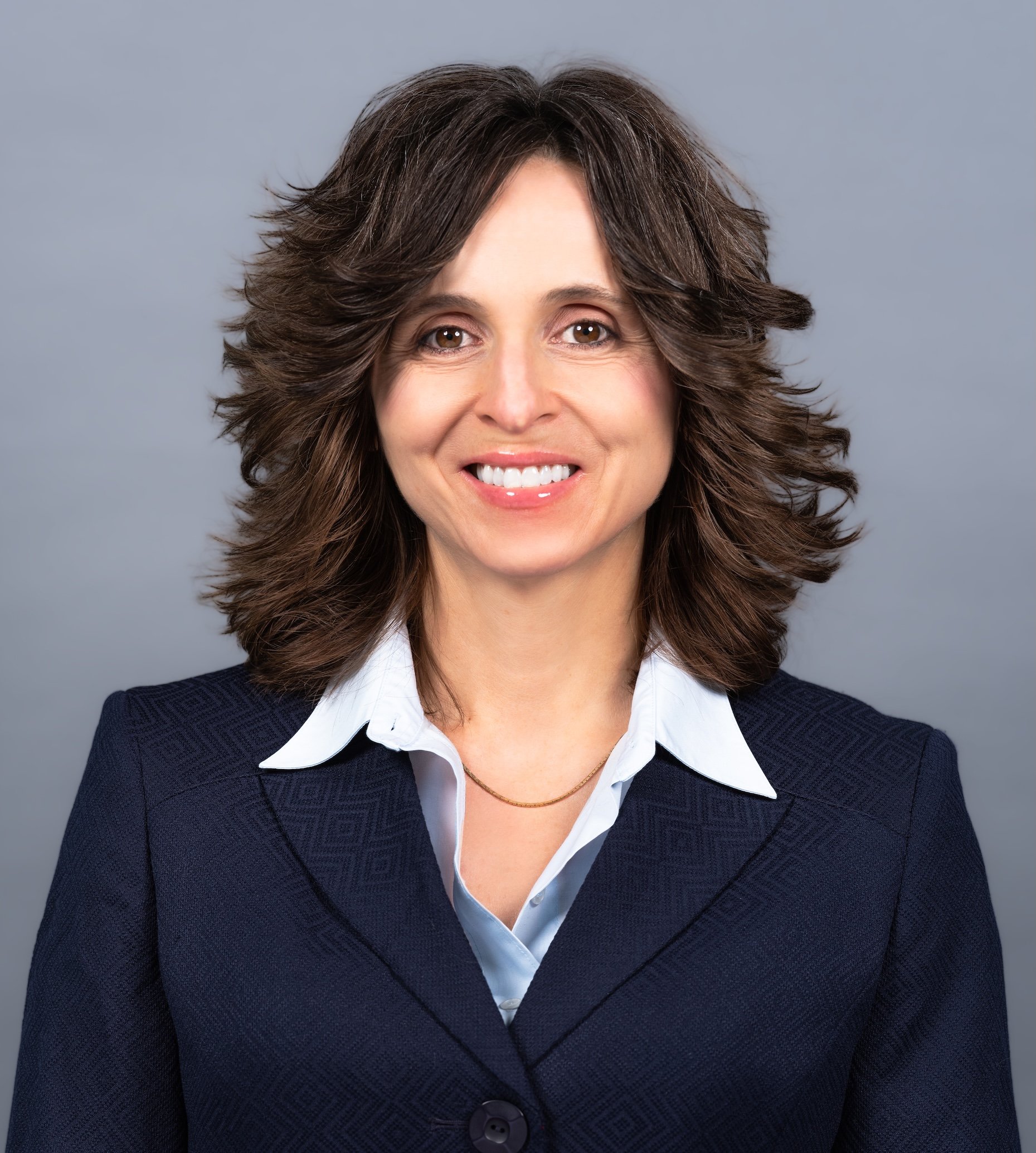 Professional Corporate Headshot by Twin Cities Photographer Jay Cupcake. Professional headshot of a woman with shoulder-length curly brown hair, wearing a dark blazer and a white collared shirt, smiling against a plain gray background.