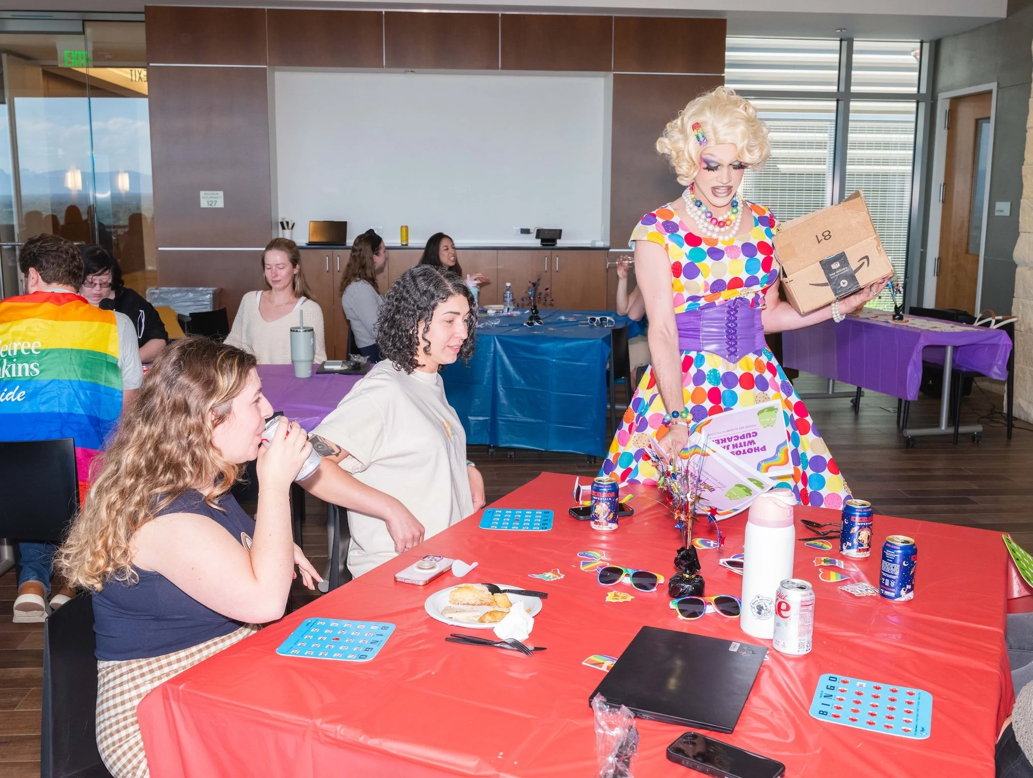 Professional Event Photography by Saint Paul Photographer Jay Cupcake. People sitting at tables decorated with rainbow and purple tablecloths during a celebration, with a person dressed in a colorful polka dot dress and wig handing out items.