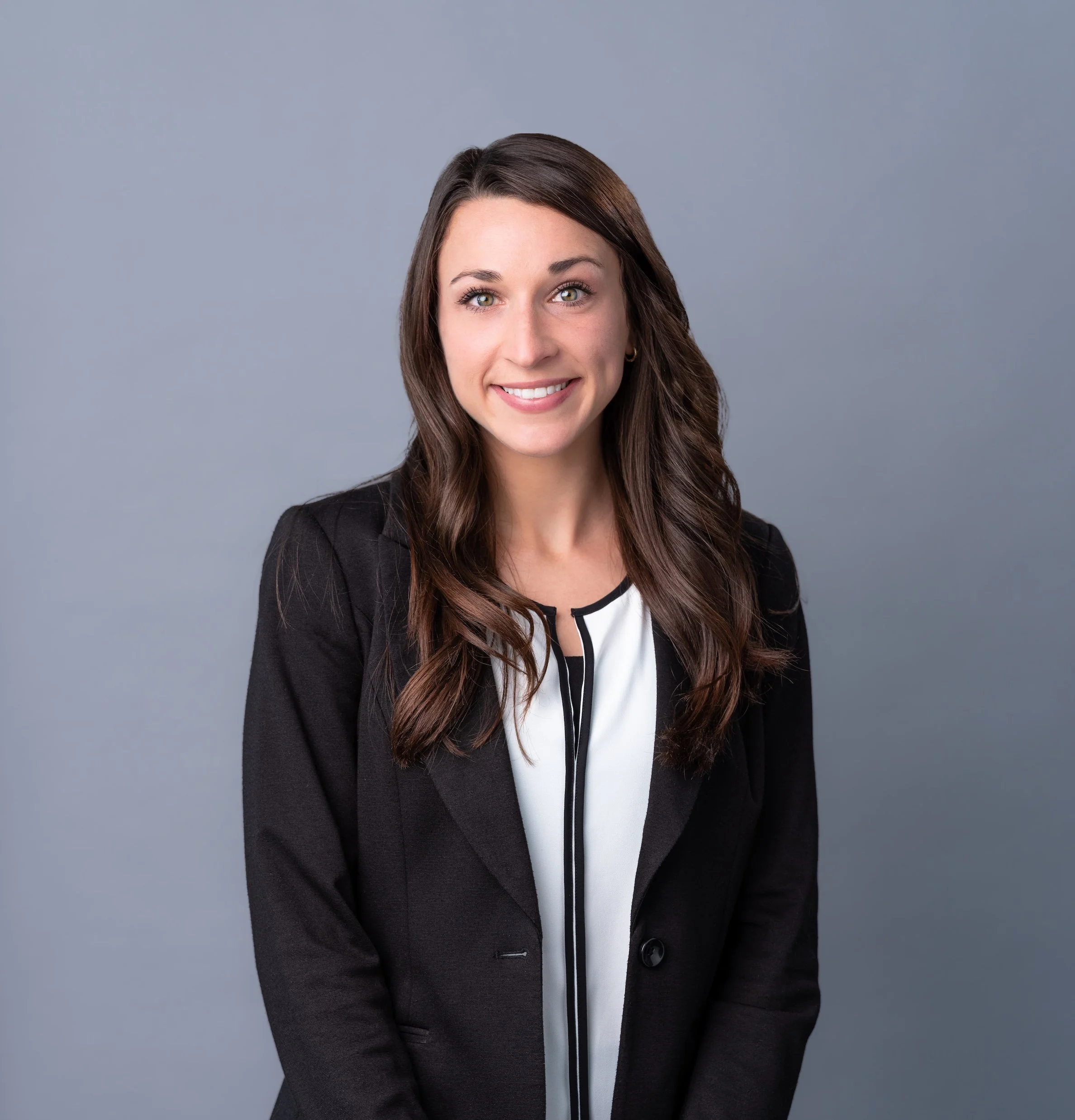 Professional Corporate Headshot by Saint Paul Photographer Jay Cupcake. Portrait of a young woman with long brown hair wearing a black blazer and white blouse, smiling against a plain gray background.