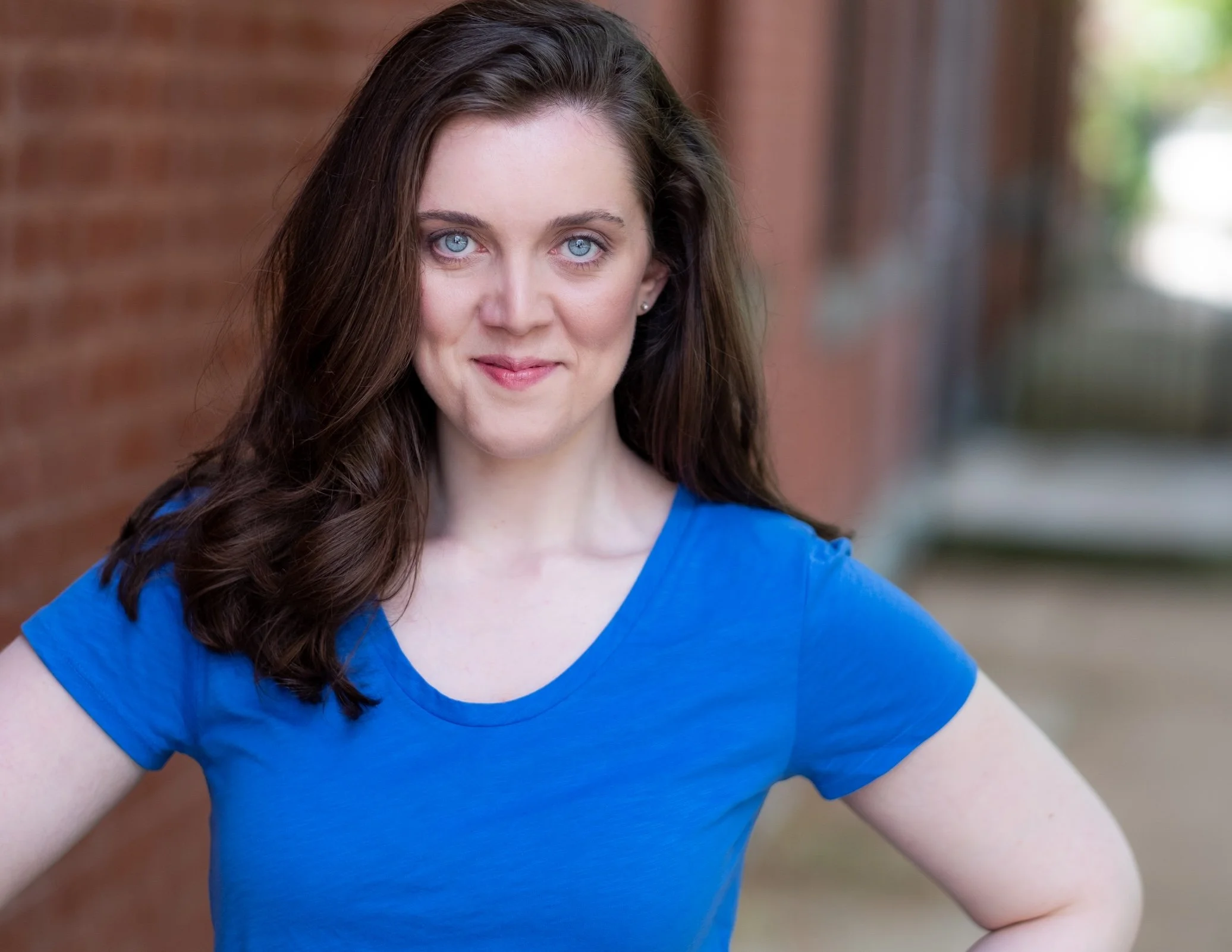 Professional Actor Headshot by Twin Cities Photographer Jay Cupcake. A woman with long brown hair and blue eyes wearing a blue t-shirt, standing outdoors near a brick wall.