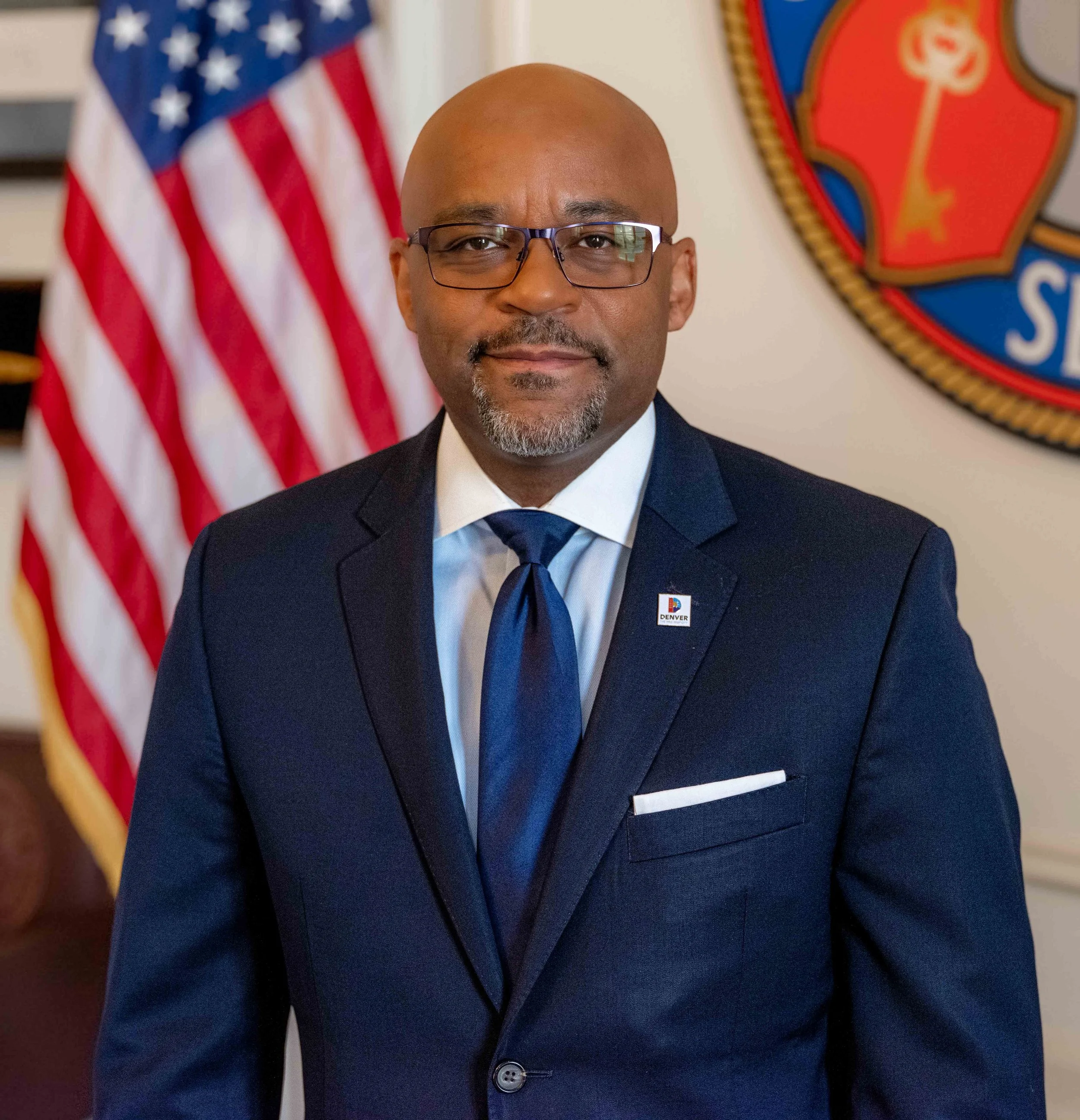Professional headshot by St. Paul photographer Jay Cupcake. A man in a navy suit, white shirt, and blue tie, wearing glasses and standing in front of an American flag and an official emblem.