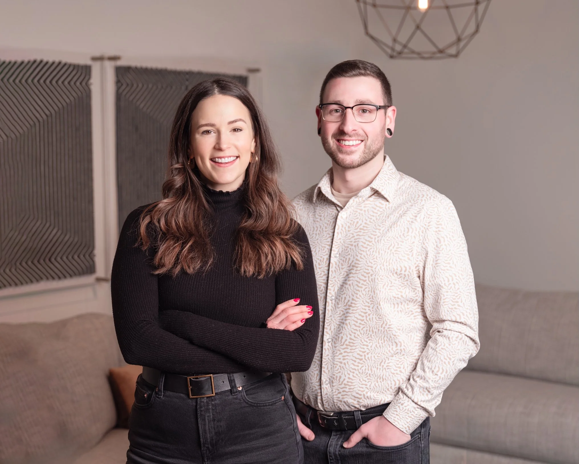 Professional Portrait Photography by Saint Paul Photographer Jay Cupcake. A man and a woman standing side by side, smiling, in a living room with beige walls, a patterned sectional sofa, and modern wall decor.
