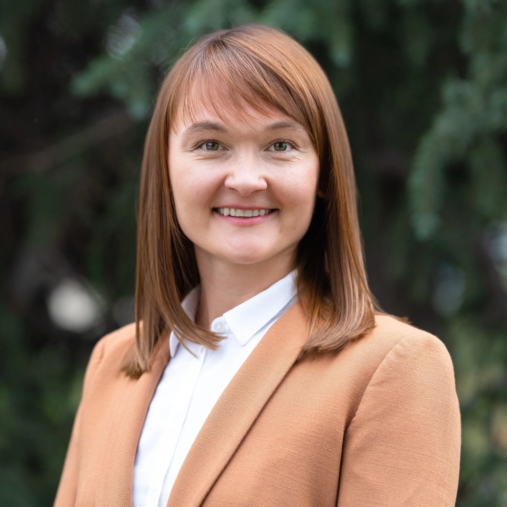 Professional Corporate Headshot by Twin Cities Photographer Jay Cupcake. A woman with shoulder-length reddish-brown hair, wearing a white shirt and tan blazer, smiling outdoors with a blurred green foliage background.
