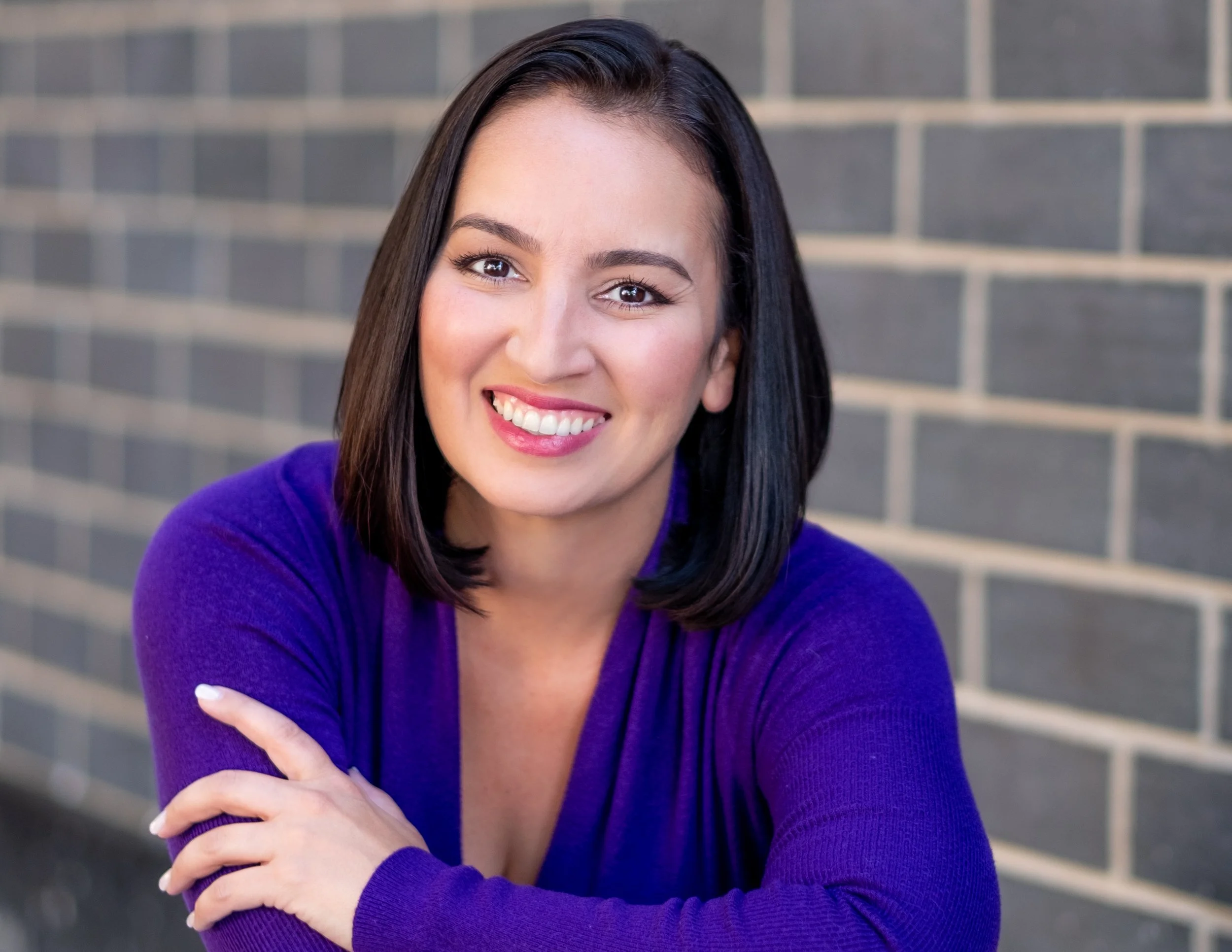 Professional headshot by St. Paul photographer Jay Cupcake. A woman with shoulder-length black hair wearing a purple top, smiling and looking at the camera, with a brick wall background.