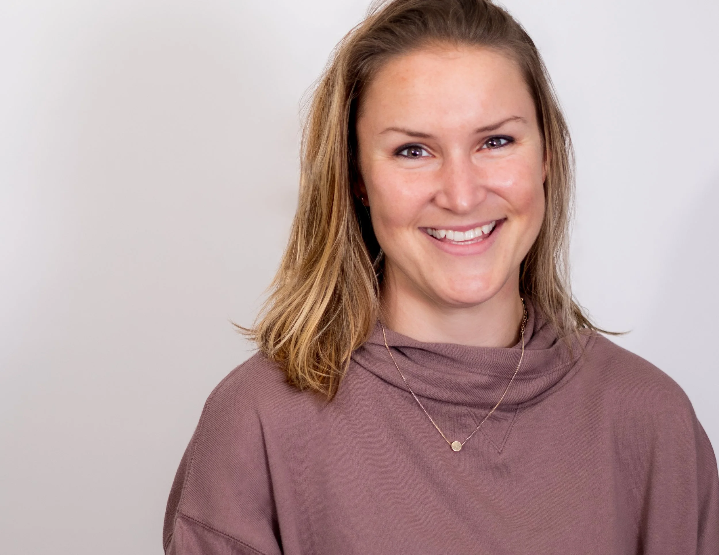 Professional headshot by St. Paul photographer Jay Cupcake. A smiling woman with shoulder-length light brown hair, wearing a mauve hoodie and a gold necklace, standing against a plain white background.