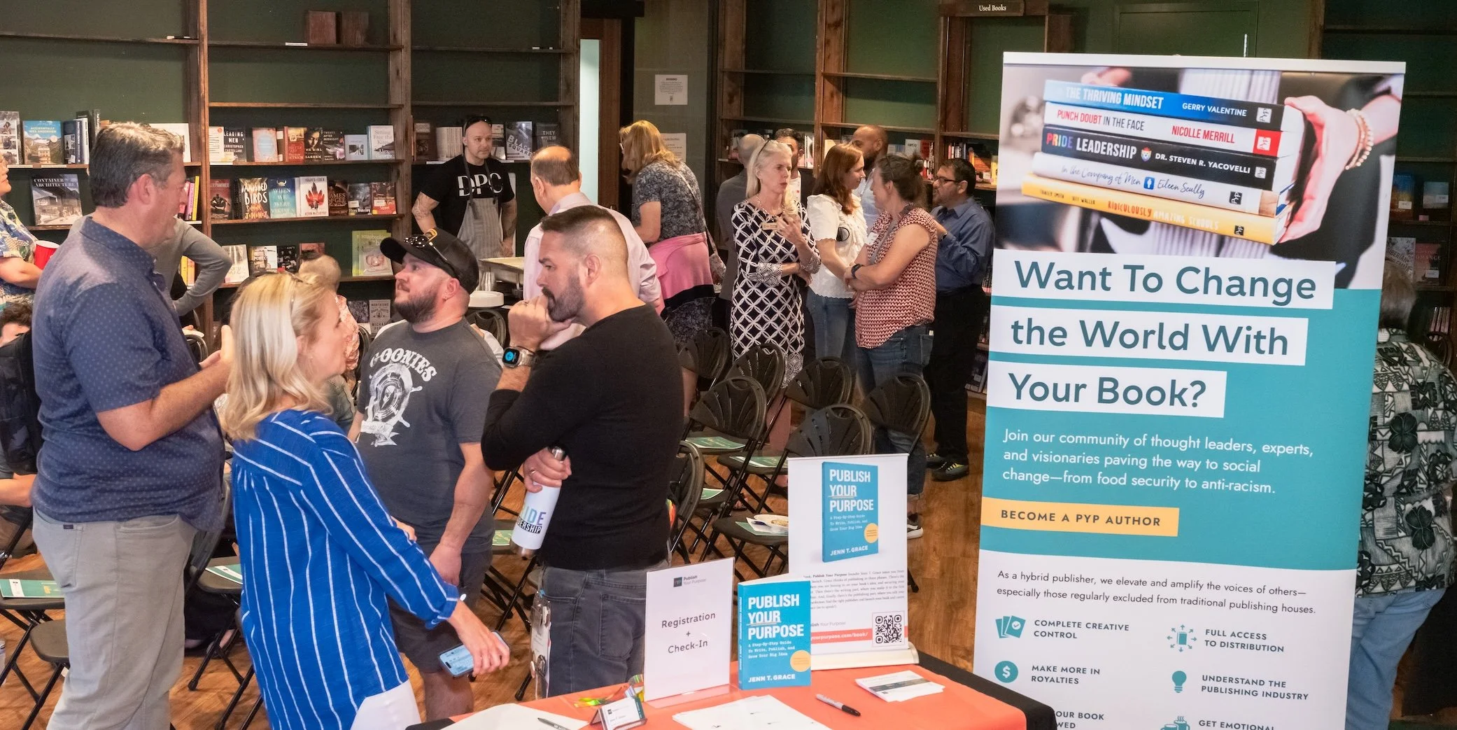 Professional Event Photography by Minneapolis Photographer Jay Cupcake. A book event or conference taking place in a bookstore or library with people engaging in conversations. A large promotional poster advertises writing and publishing books to cha