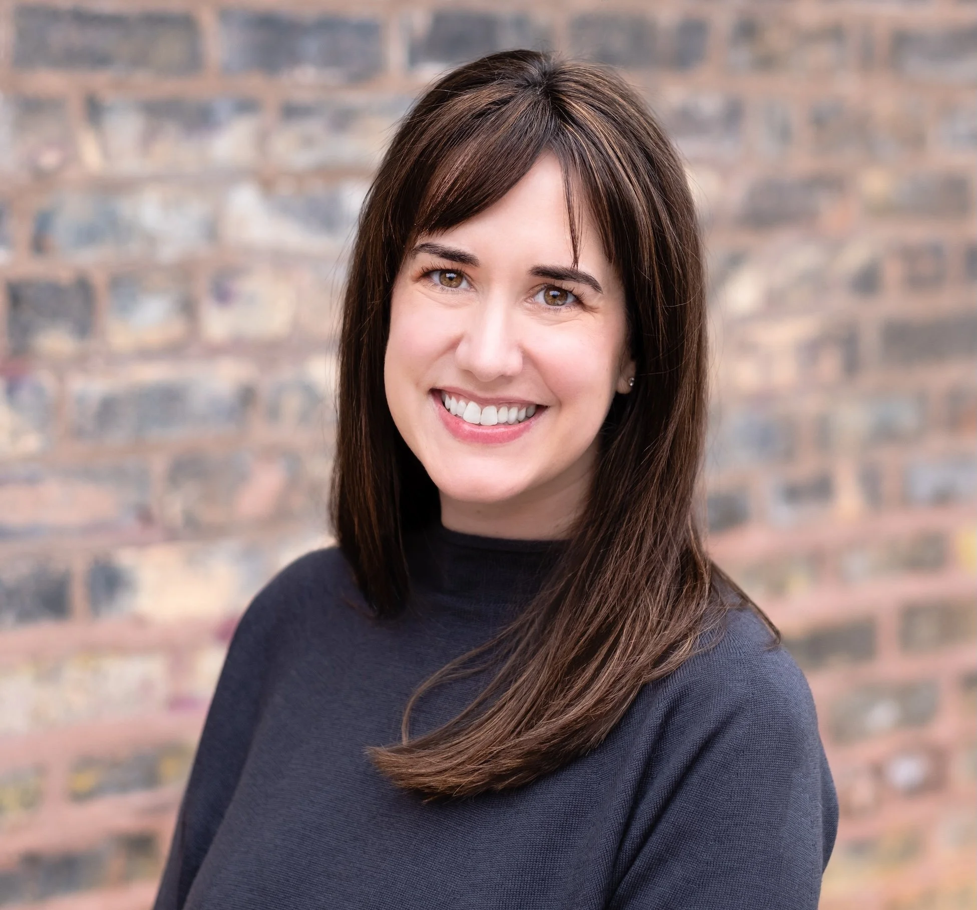Professional Corporate Headshot by Minneapolis Photographer Jay Cupcake. A smiling woman with straight brown hair and brown eyes, wearing a black top, standing against a brick wall background.