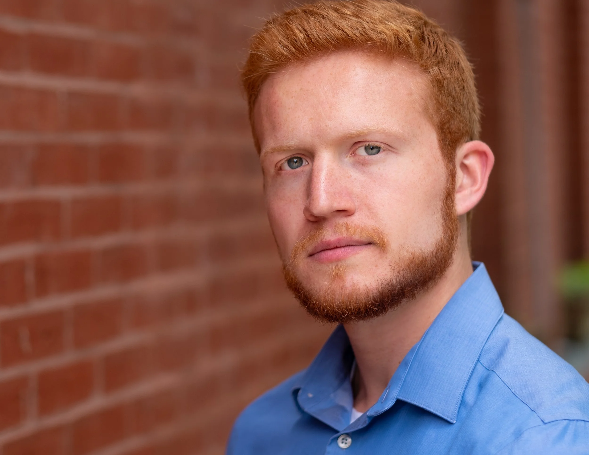 Professional headshot by St. Paul photographer Jay Cupcake. A young man with red hair, blue eyes, and a beard, wearing a blue shirt, standing outdoors next to a red brick wall.