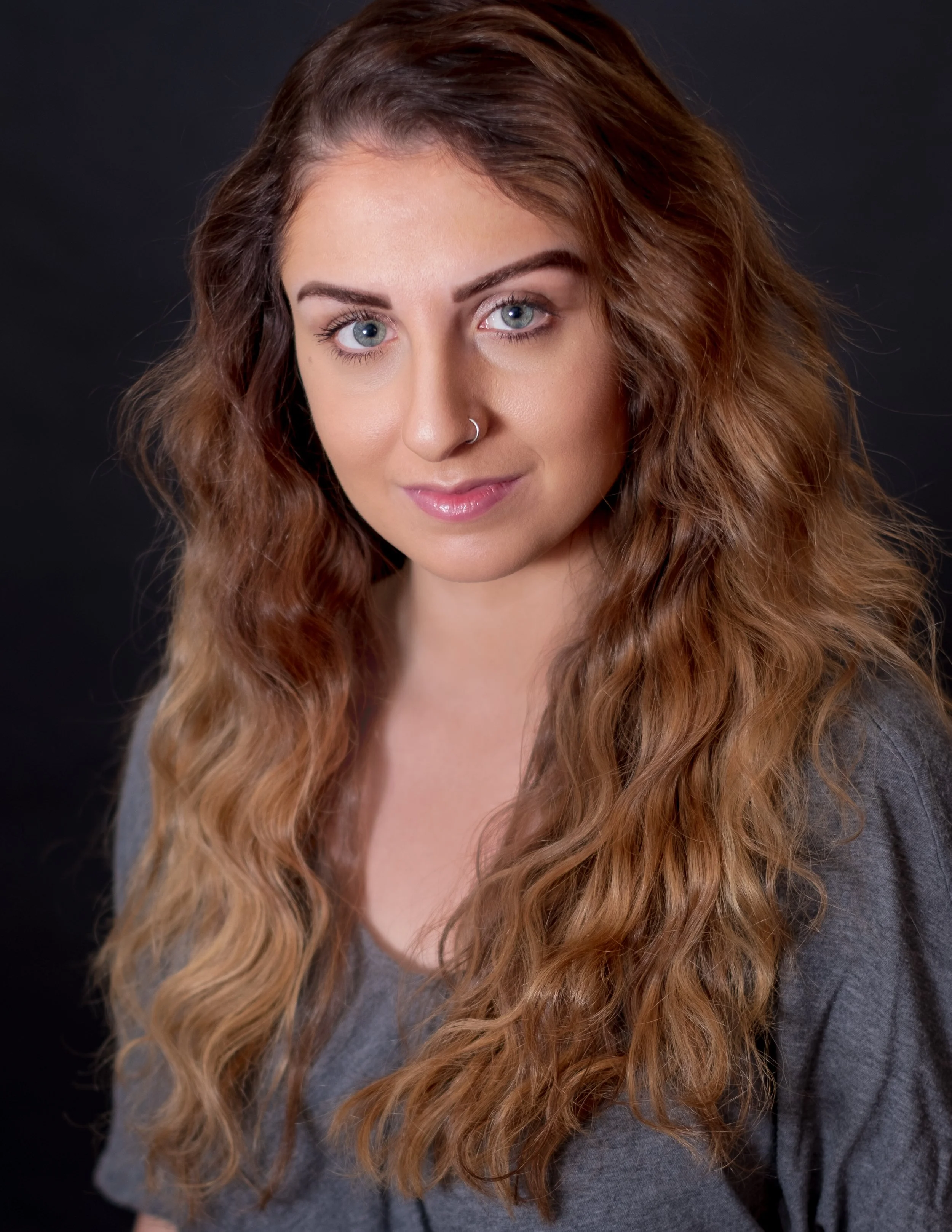Professional headshot by St. Paul photographer Jay Cupcake. Portrait of a young woman with blue eyes, light makeup, wavy auburn hair, wearing a gray top, against a dark background.