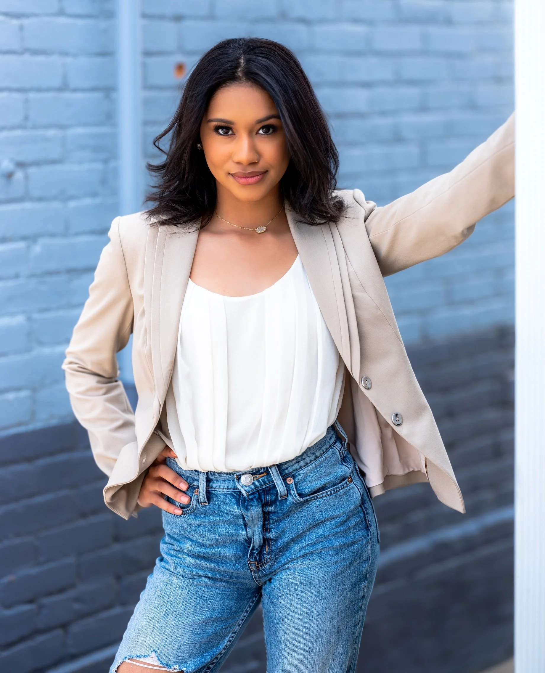 Professional Portrait Photography by Saint Paul Photographer Jay Cupcake. A young woman with dark, shoulder-length hair, wearing a beige blazer over a white blouse, paired with ripped blue jeans, standing against a blue brick wall, looking confidentl