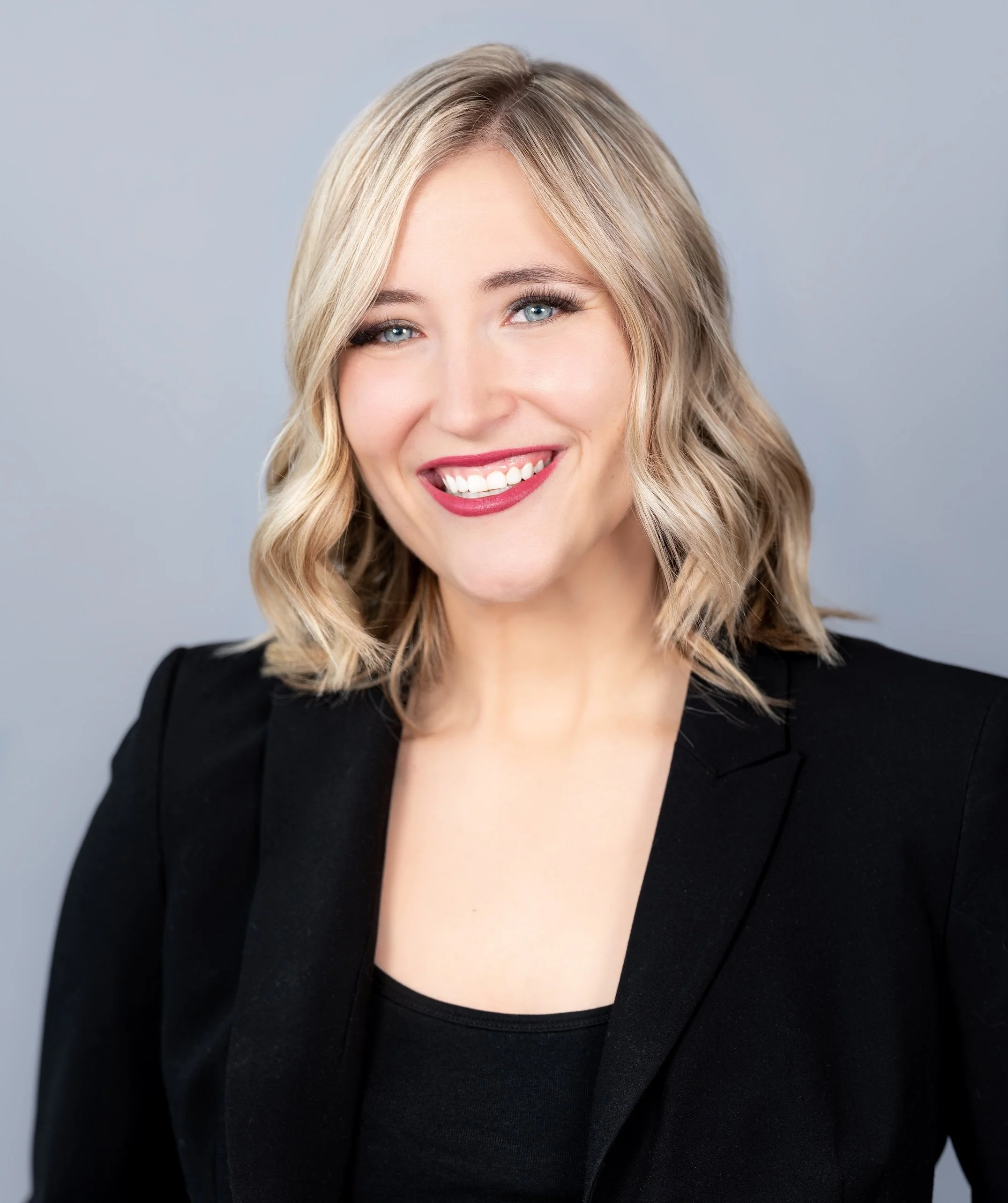 Professional Corporate Headshot by Saint Paul Photographer Jay Cupcake. A smiling woman with blonde hair styled in loose waves, wearing a black blazer over a black top, against a light gray background.