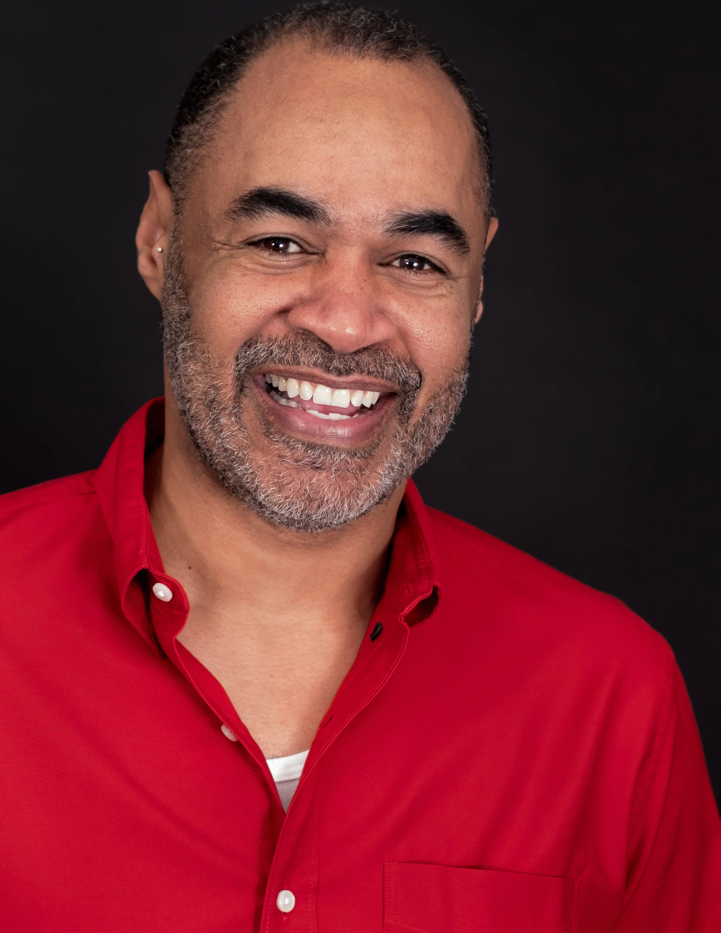 Professional headshot by St. Paul photographer Jay Cupcake. A smiling man with dark curly hair, a beard with gray and black hair, wearing a red shirt, against a dark background.