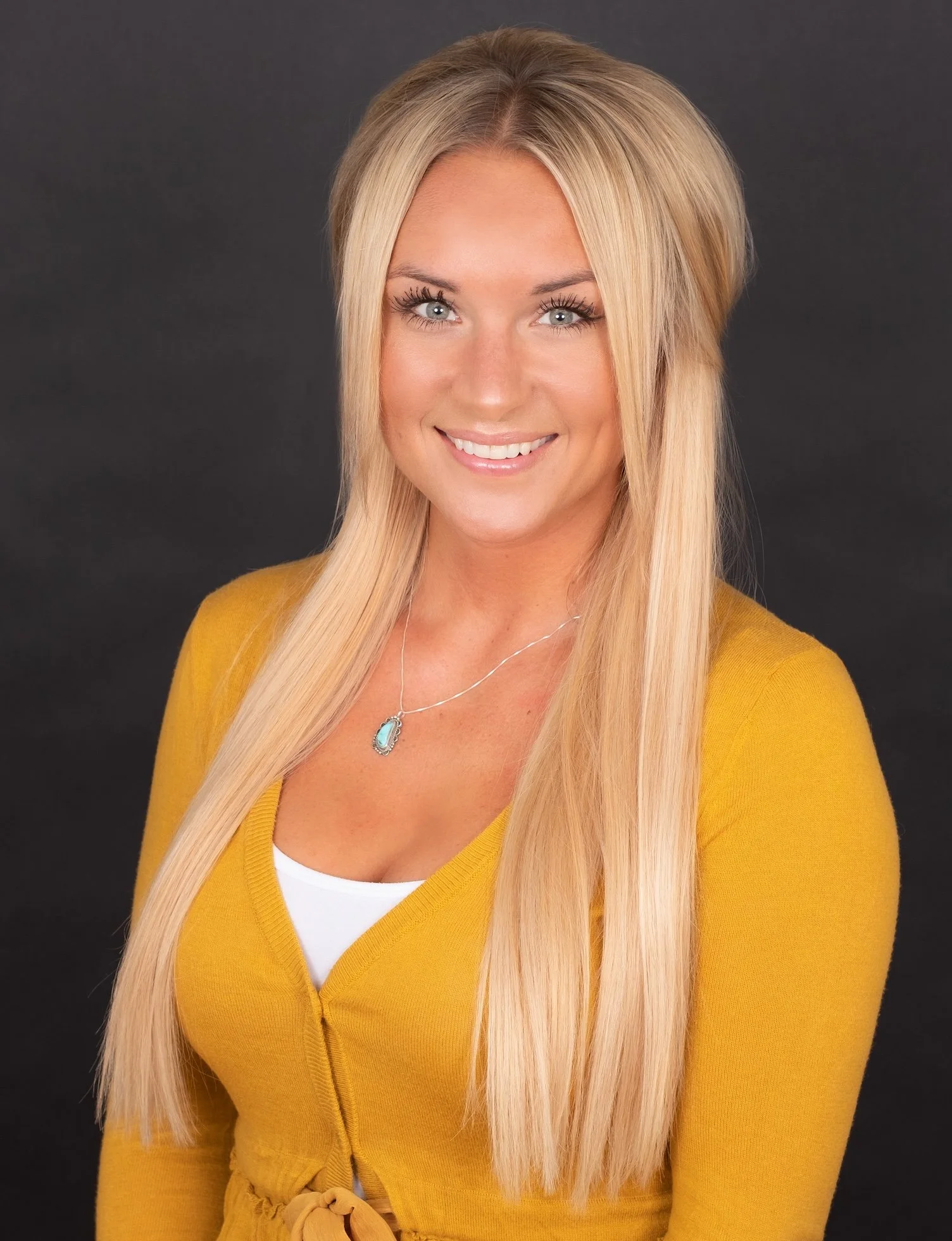 Professional Corporate Headshot by Twin Cities Photographer Jay Cupcake. A woman with long blonde hair wearing a yellow cardigan and a turquoise pendant necklace, smiling facing the camera against a dark background.