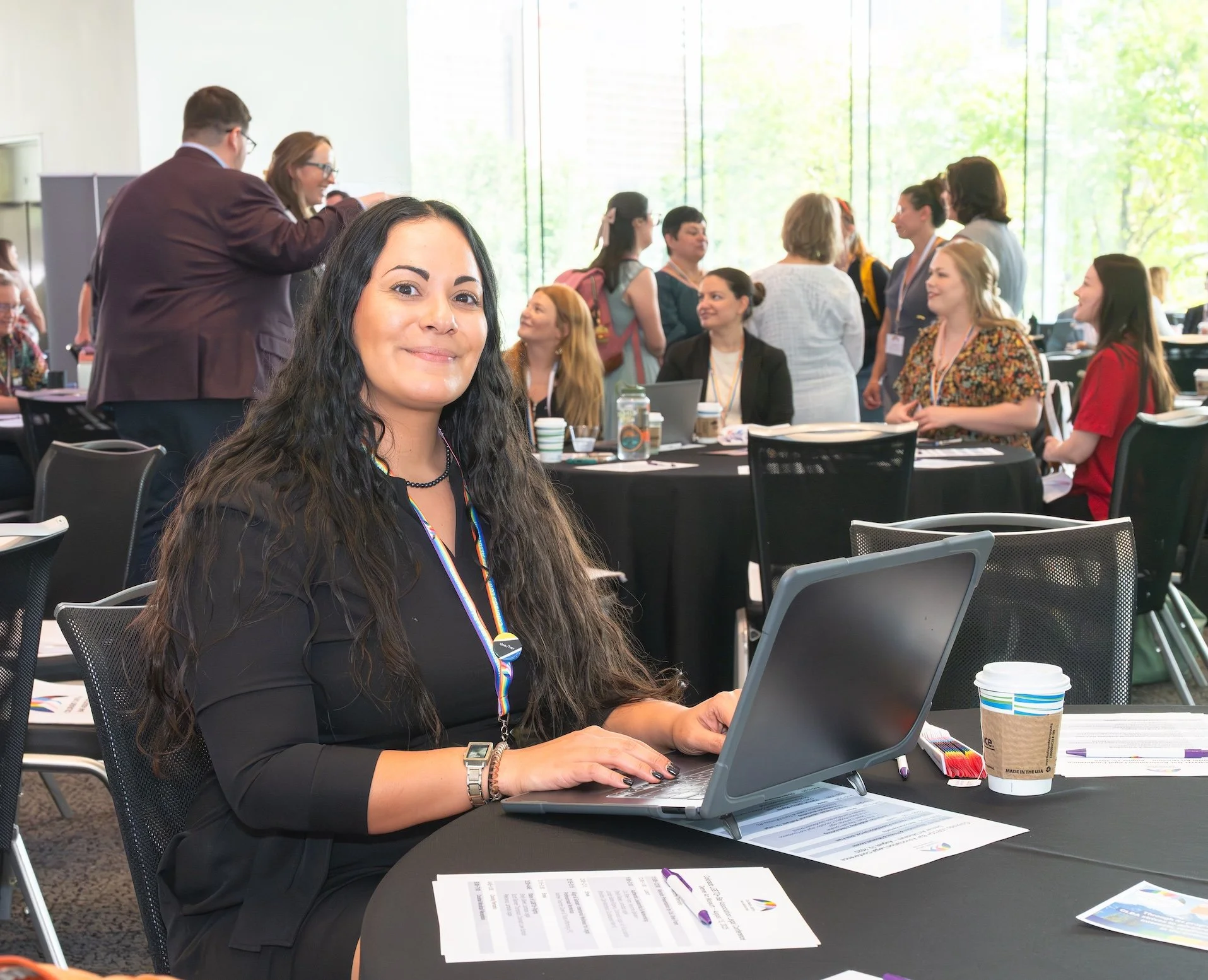 Professional Event Photography by Saint Paul Photographer Jay Cupcake. Woman with long dark hair smiling at camera while working on a laptop at a conference table in a busy conference room with other attendees in the background.