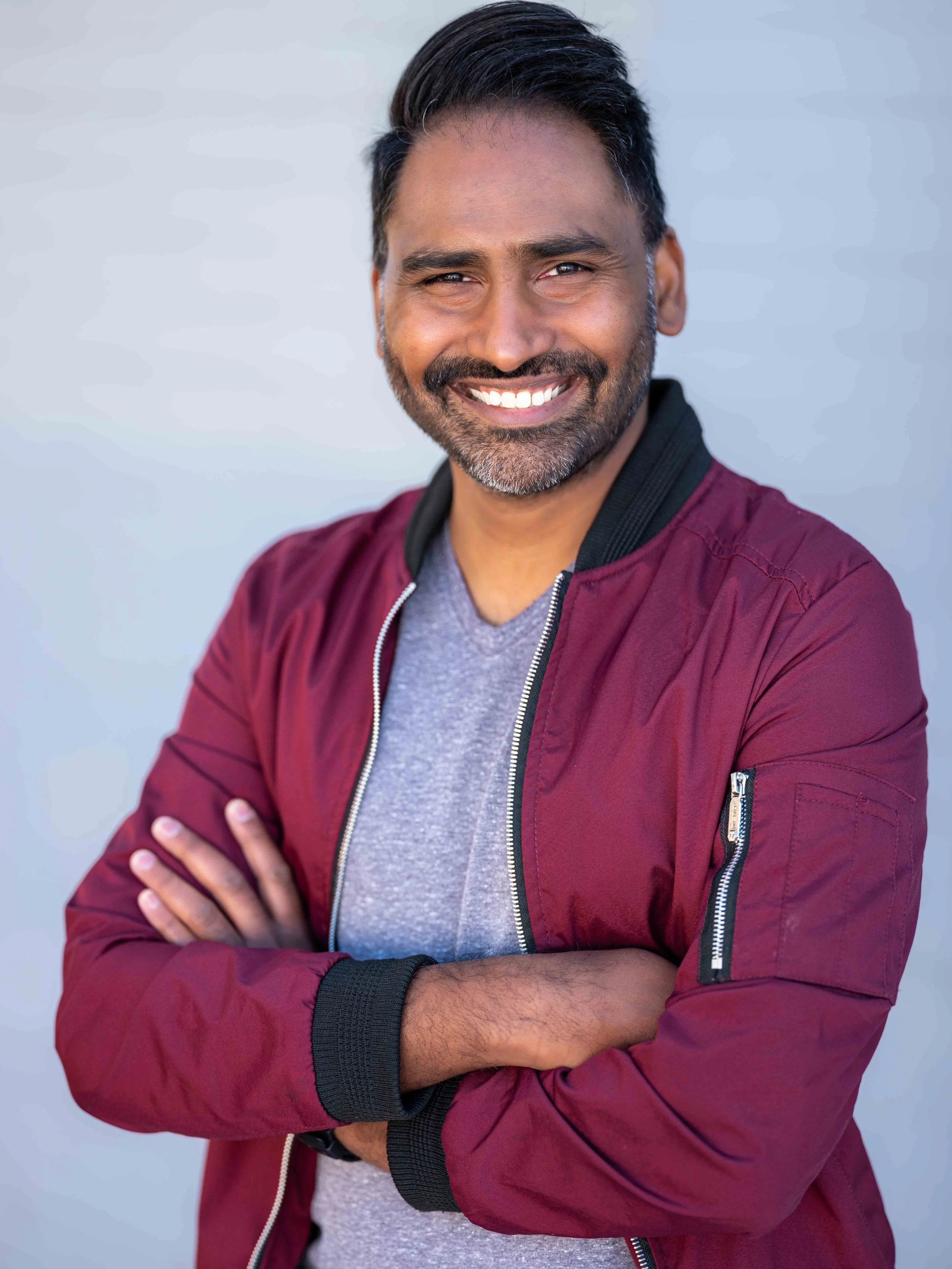 Professional headshot by St. Paul photographer Jay Cupcake. A smiling man with dark hair, beard, and mustache, wearing a maroon jackets and a gray shirt, with arms crossed, standing against a plain gray background.