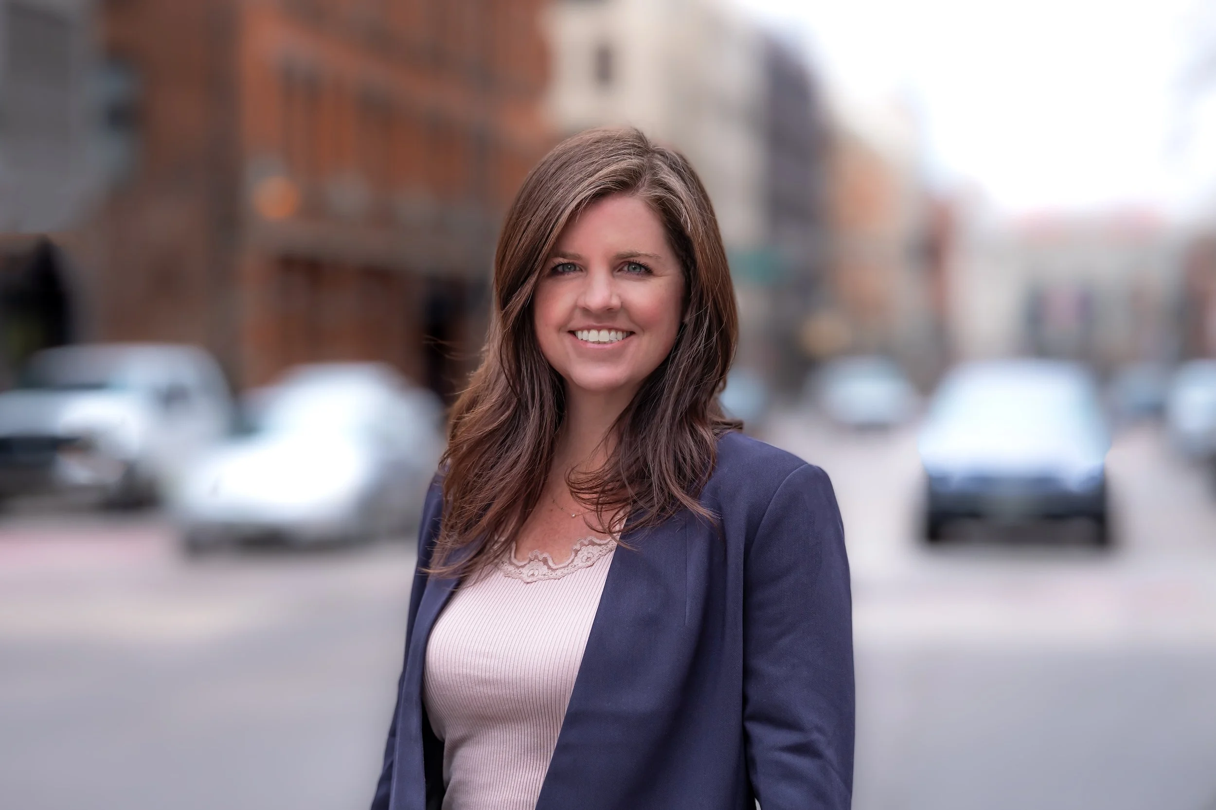 Professional Corporate Headshot by Twin Cities Photographer Jay Cupcake. A woman with long brown hair smiling, standing outdoors in front of a blurred street scene with parked cars and brick buildings.