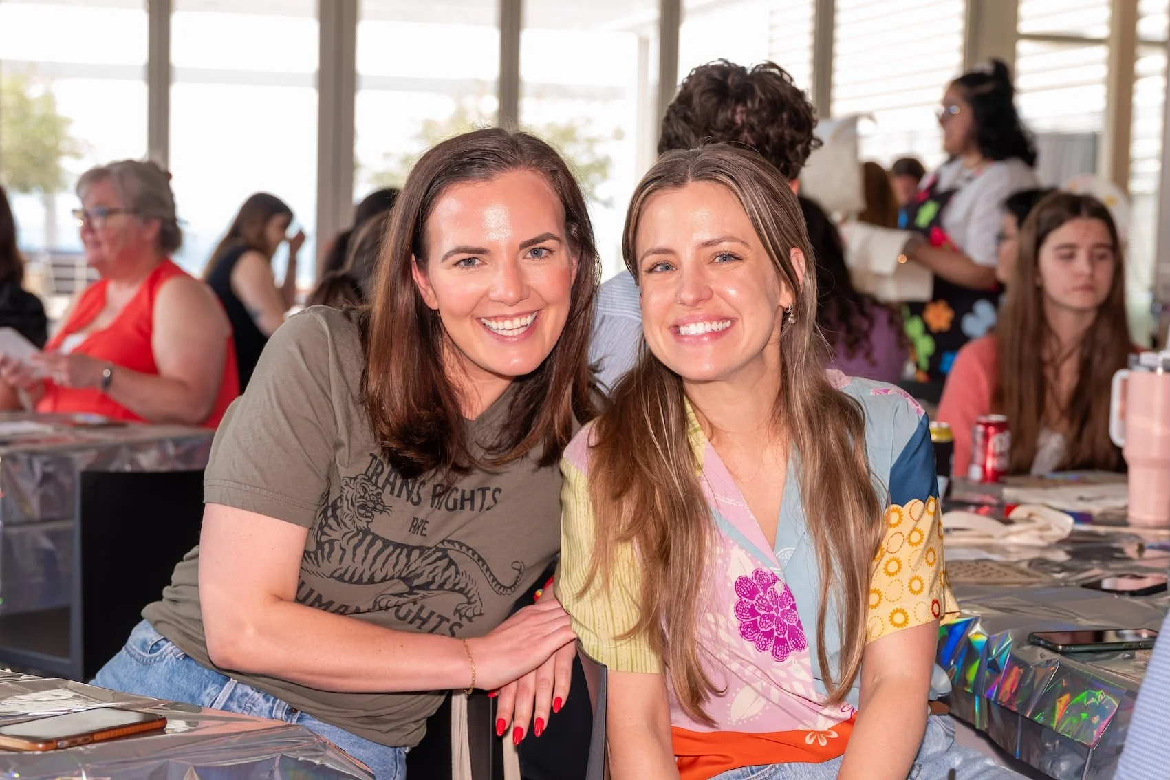 Professional Event Photography by Twin Cities Photographer Jay Cupcake. Two women smiling and posing together at a gathering, with other people seated at tables in the background.