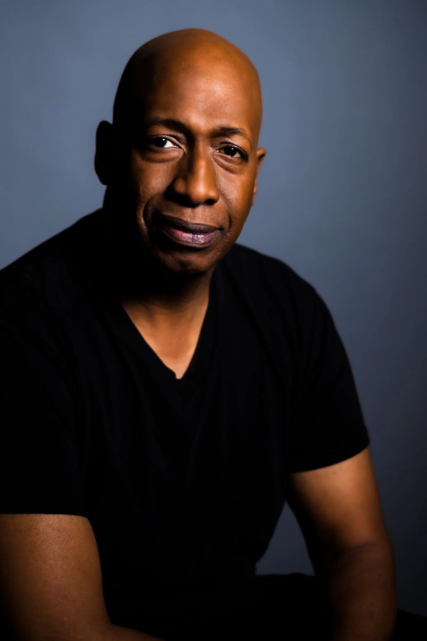 Professional headshot by St. Paul photographer Jay Cupcake. A portrait of a bald African American man wearing a black shirt, looking at the camera with a serious expression against a dark background.