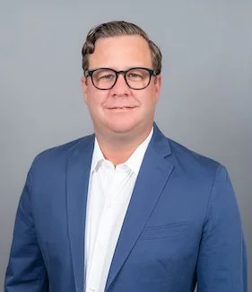 Professional Corporate Headshot by Twin Cities Photographer Jay Cupcake. A man with glasses wearing a blue suit and white shirt, standing against a gray background.