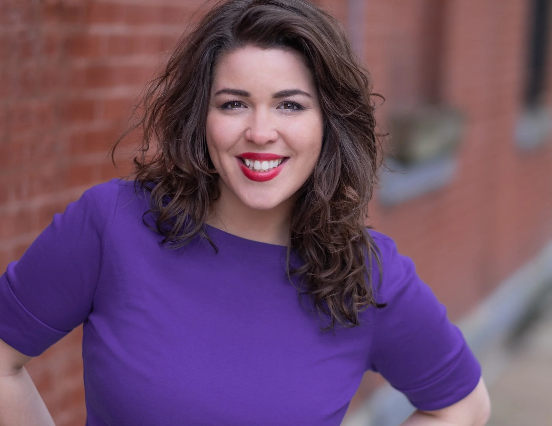 Professional Actor Headshot by Saint Paul Photographer Jay Cupcake. A woman with curly brown hair and red lipstick smiling, wearing a purple shirt, standing outdoors in front of a brick wall.