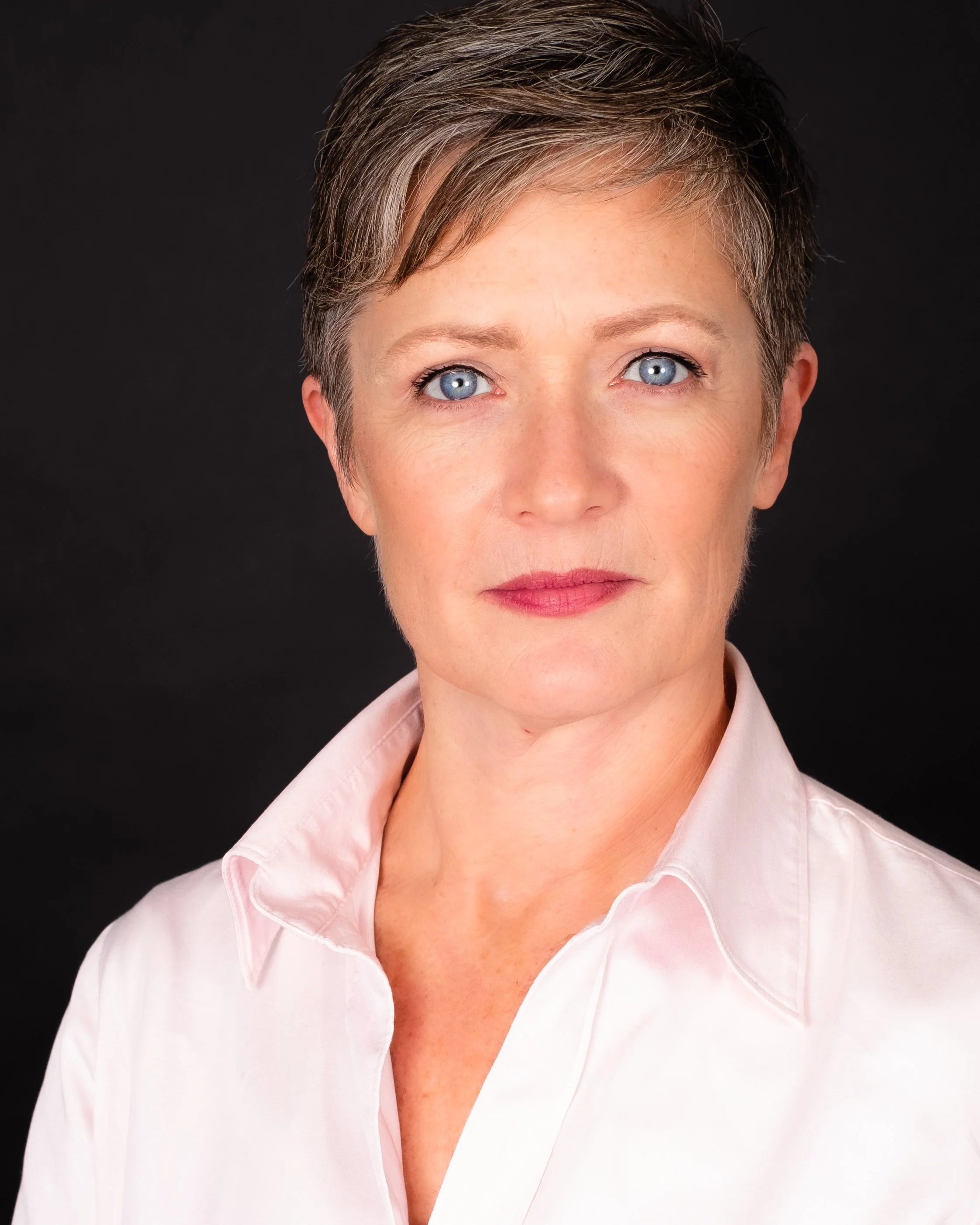 Professional headshot by St. Paul photographer Jay Cupcake. A woman with short gray hair and blue eyes wearing a white shirt against a black background.