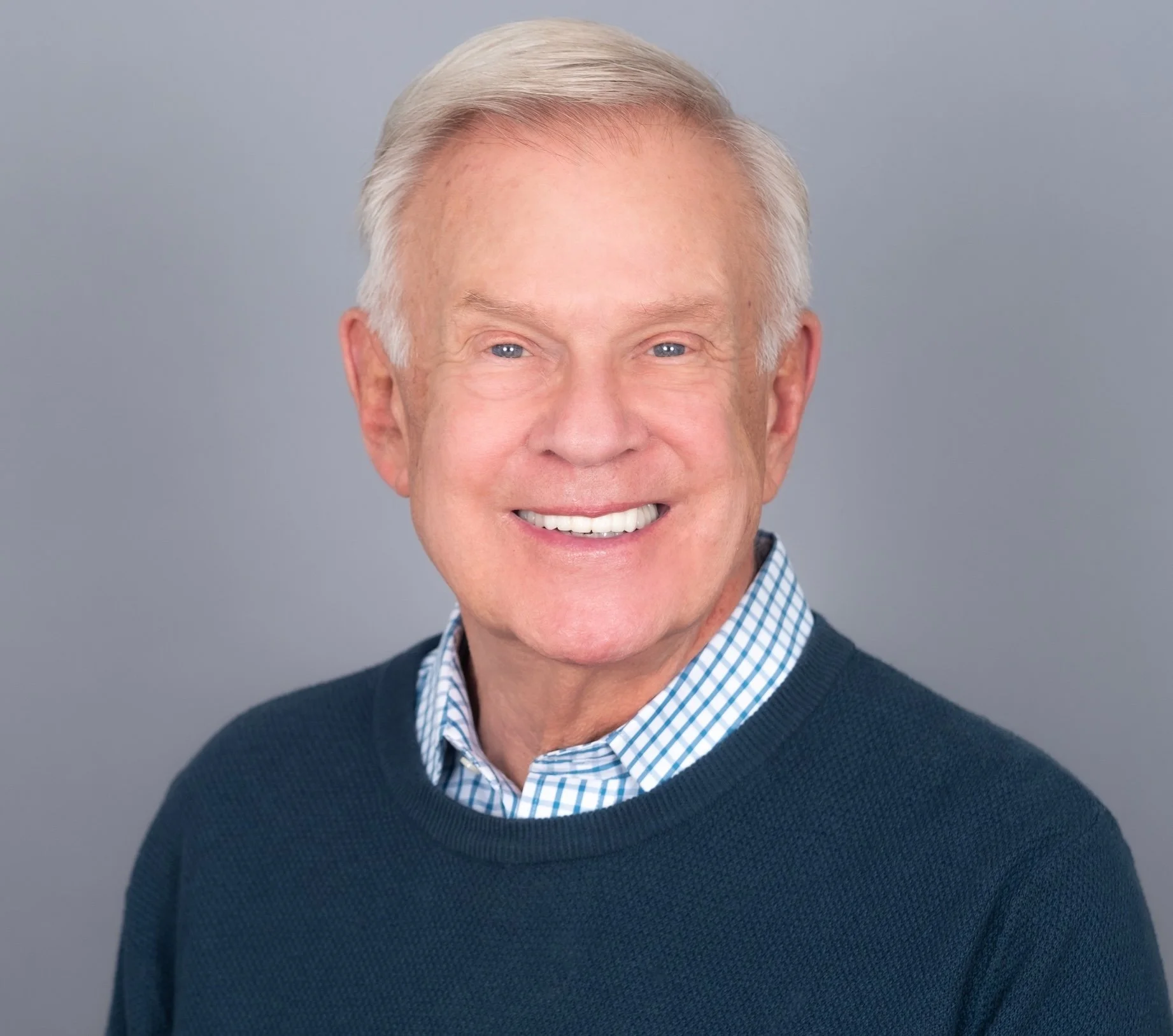 Professional Corporate Headshot by Saint Paul Photographer Jay Cupcake. Portrait of an elderly man with short, white hair, smiling, wearing a blue sweater over a checkered shirt, against a gray background.