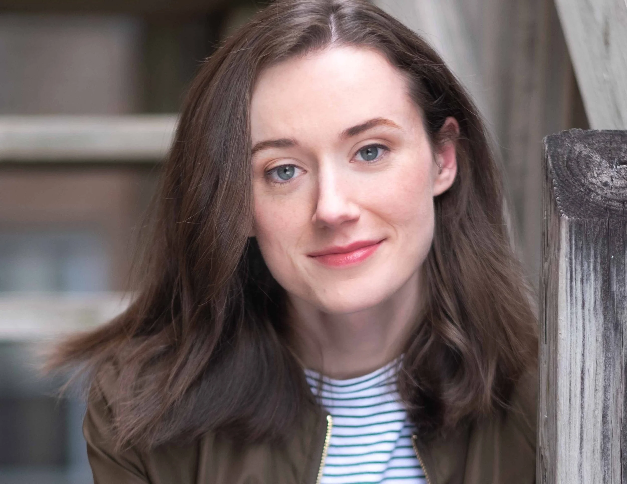 Professional Actor Headshot by Saint Paul Photographer Jay Cupcake. Close-up of a young woman with shoulder-length brown hair, blue eyes, and light skin, smiling outdoors near a wooden structure, wearing a striped shirt and a brown jacket.