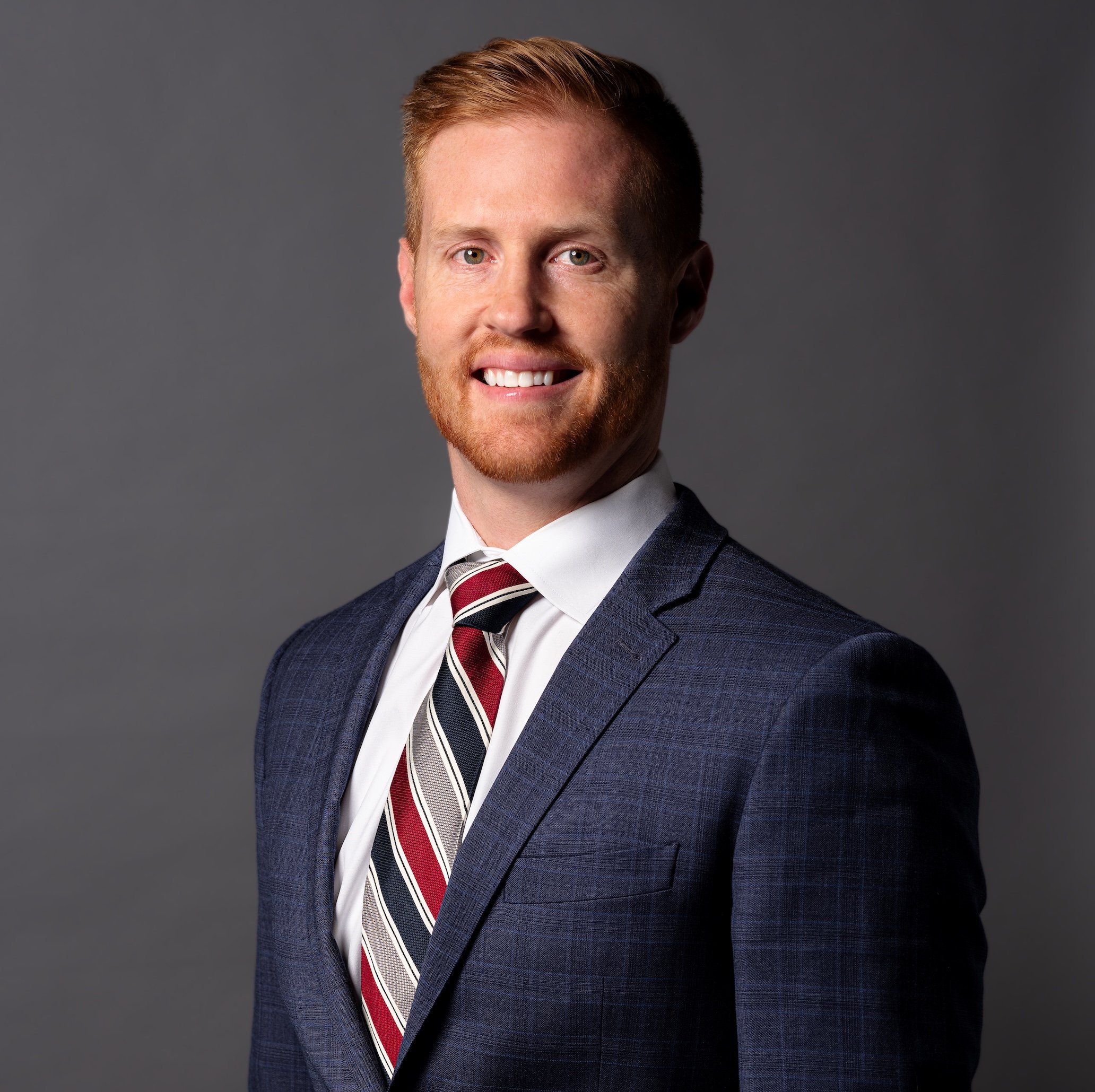 Professional Corporate Headshot by Minneapolis Photographer Jay Cupcake. A portrait of a young man with red hair, a beard, wearing a navy blue suit, white shirt, and a red, white, and blue striped tie, smiling against a gray background.