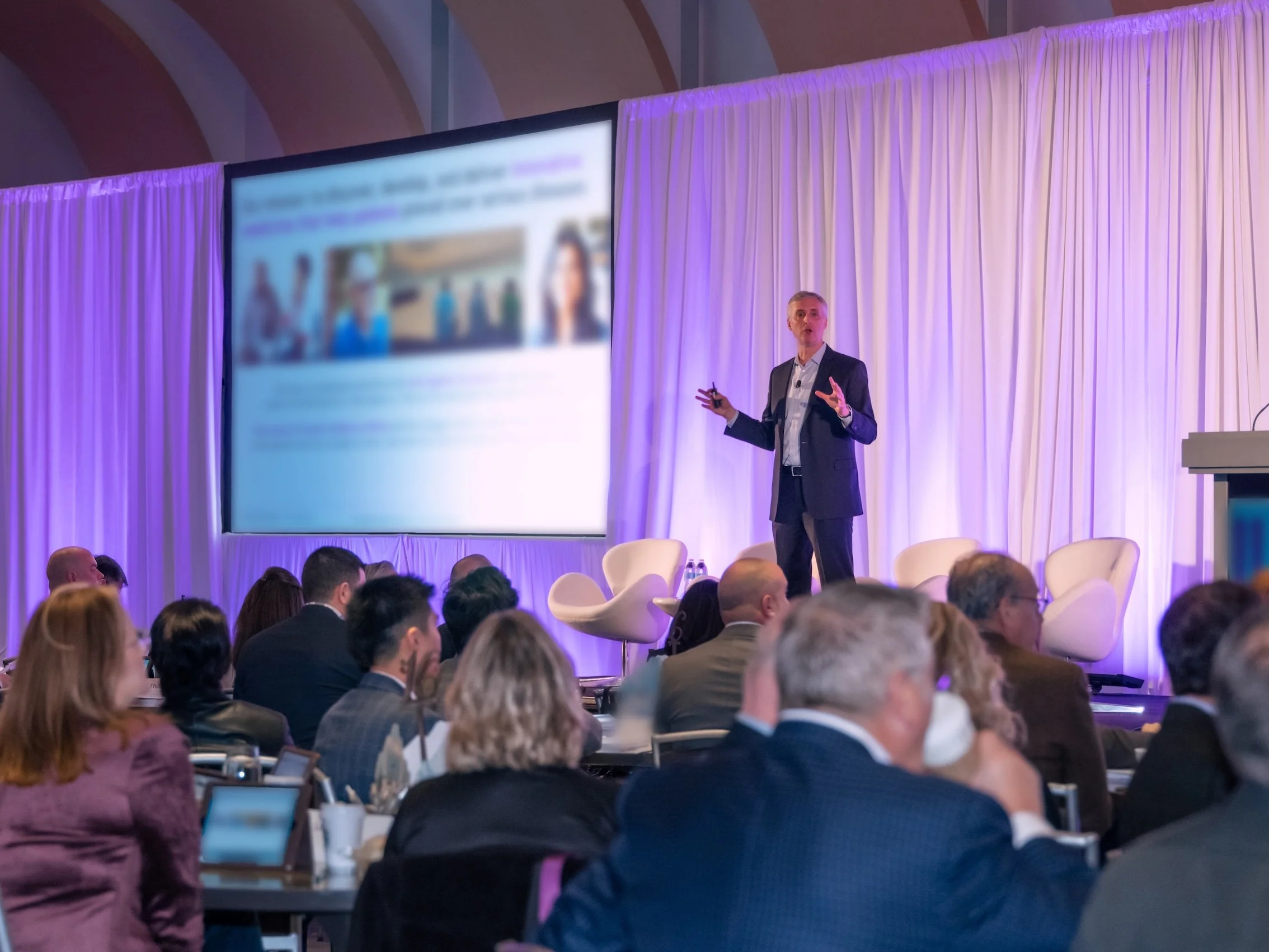 Professional Event Photography by Minneapolis Photographer Jay Cupcake. A man giving a presentation on stage at a conference, with a large blurred screen behind him. Audience members are seated, facing the stage.