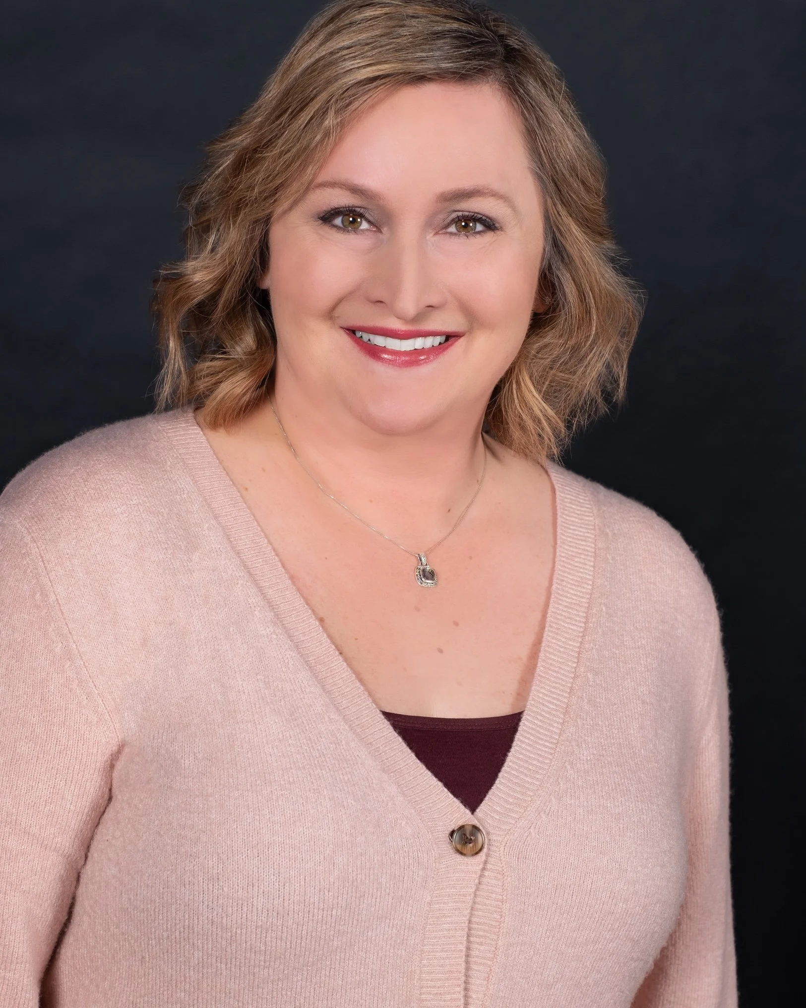 Professional Actor Headshot by Twin Cities Photographer Jay Cupcake. A woman with fair skin, shoulder-length light brown hair, and wearing a light pink cardigan over a dark top, smiling in front of a black background.