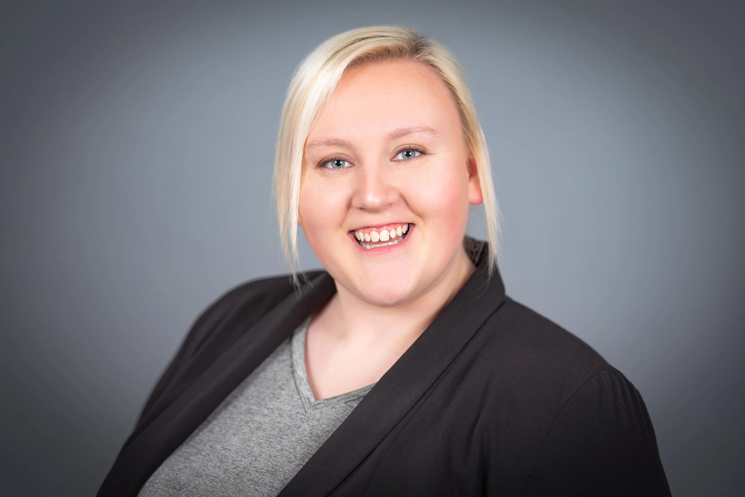 Professional Corporate Headshot by Twin Cities Photographer Jay Cupcake. Portrait of a young woman with blonde hair smiling, wearing a black blazer and gray top against a gray background.