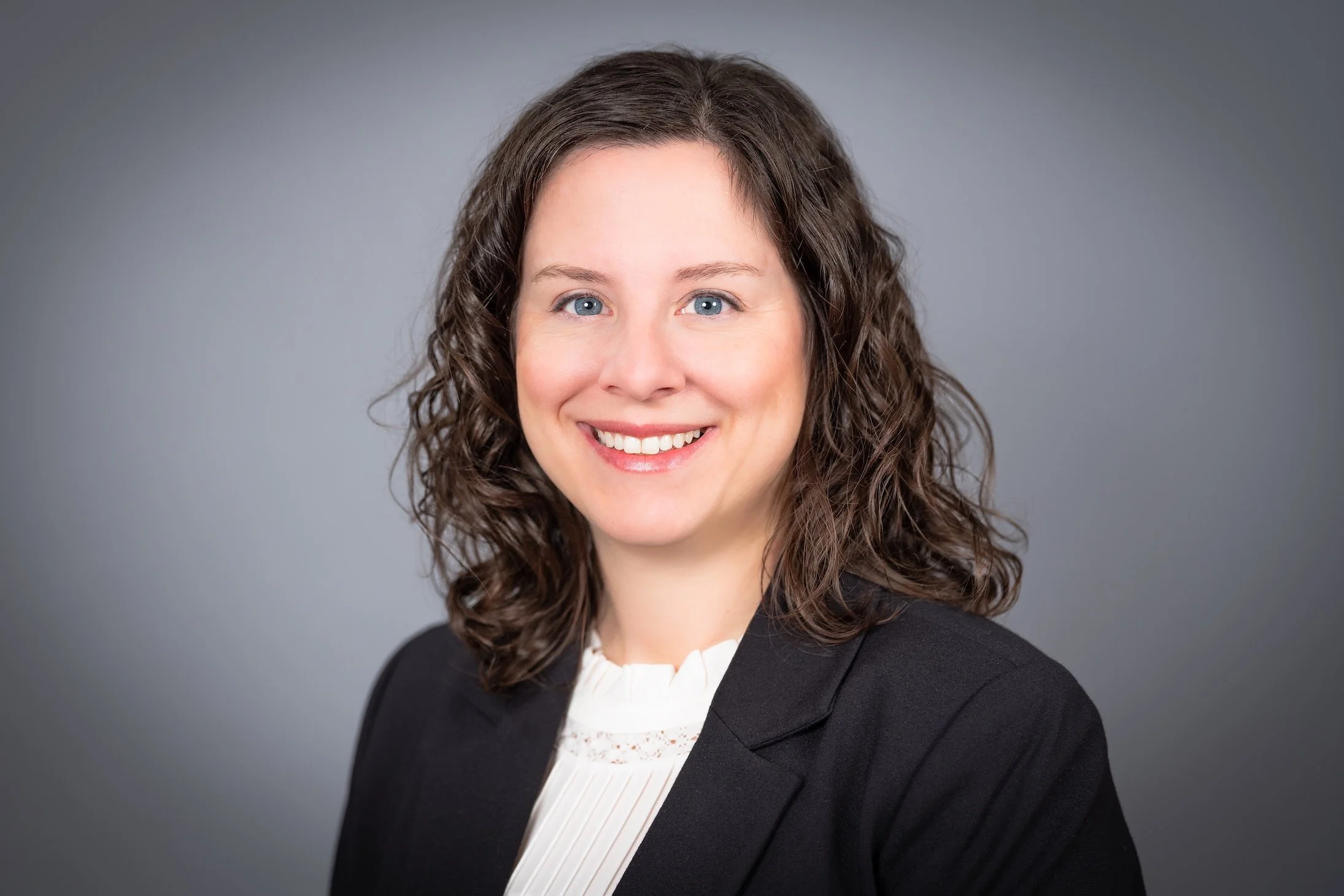 Professional Corporate Headshot by Saint Paul Photographer Jay Cupcake. Professional headshot of a woman with shoulder-length curly brown hair, wearing a black blazer and white blouse, smiling against a gray background.