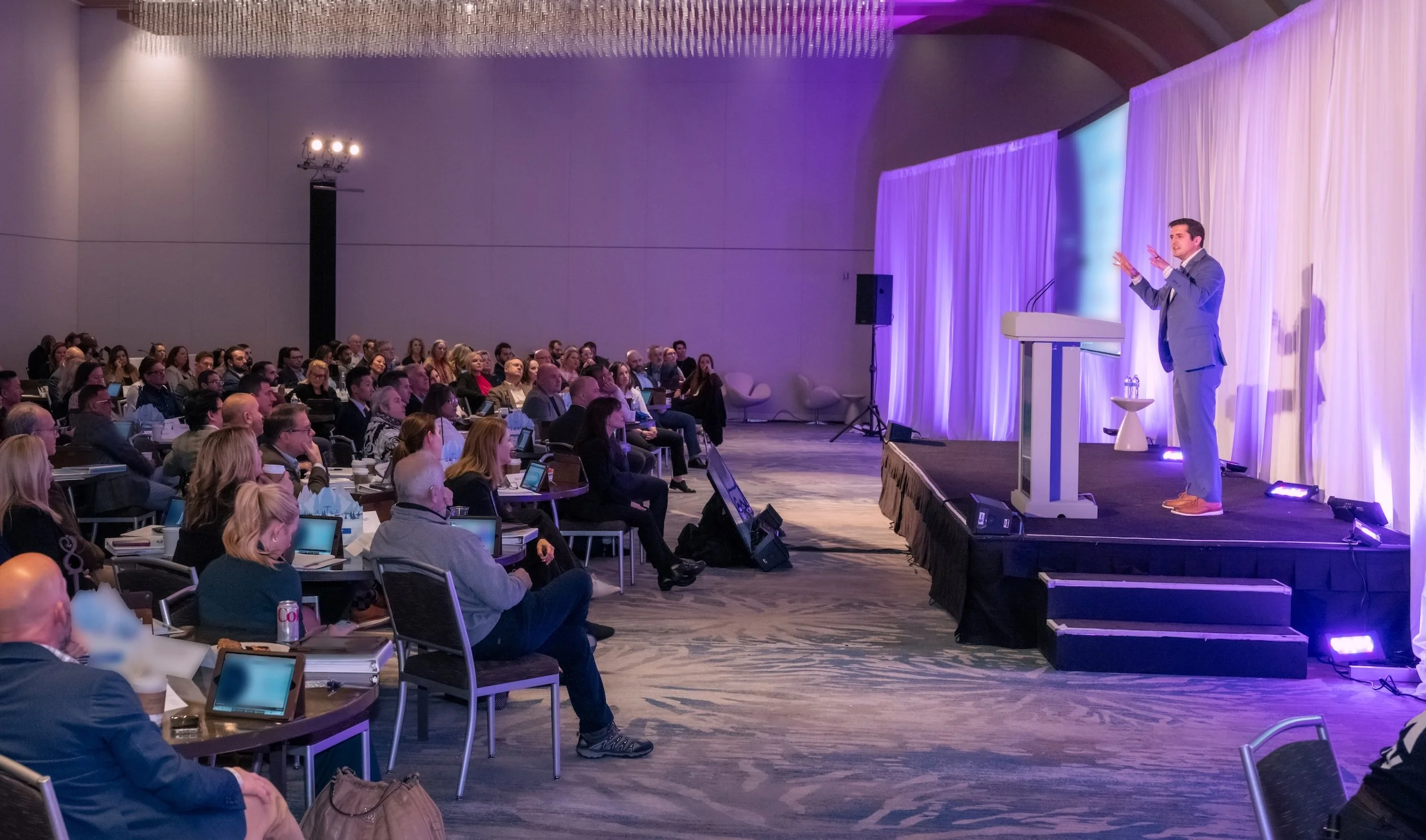 Professional Event Photography by Twin Cities Photographer Jay Cupcake. A speaker on stage delivering a presentation at a conference, with a large audience sitting at round tables watching attentively. The stage is decorated with purple lighting and 