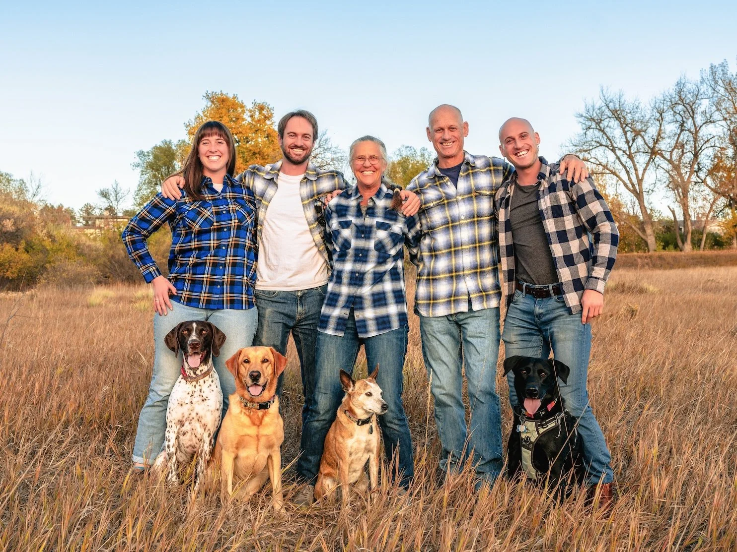 Professional Portrait Photography by Minneapolis Photographer Jay Cupcake. Group of five people and four dogs standing in a grassy field during autumn, smiling with trees and a blue sky in the background.