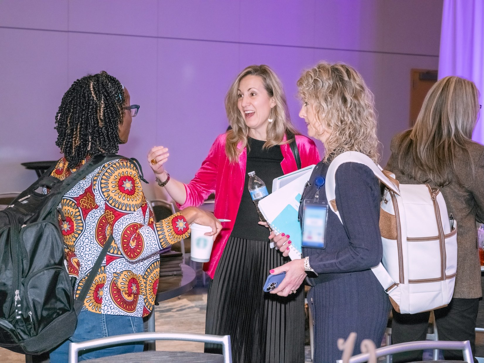 Professional Event Photography by Saint Paul Photographer Jay Cupcake. Four women engaged in conversation at a professional event, one woman wearing a patterned jacket and carrying a backpack, another with a water bottle, and another with a large tot