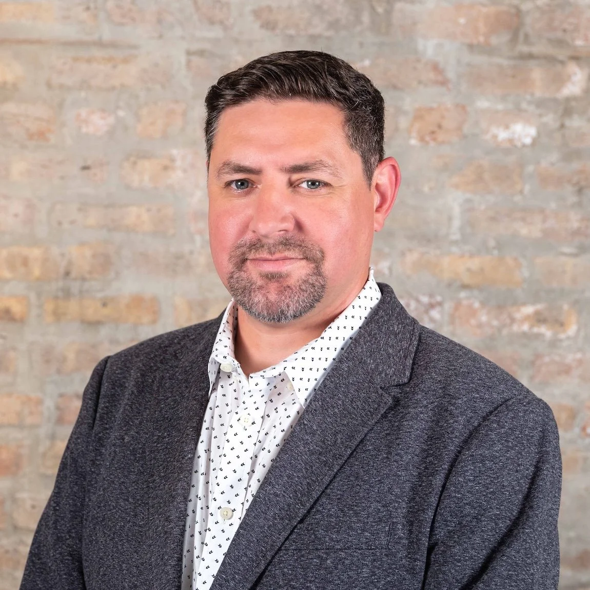 Professional Corporate Headshot by Twin Cities Photographer Jay Cupcake. A man with short dark hair, a beard, and light skin wearing a dark gray blazer and a white shirt with small black patterns, standing against a brick wall background.