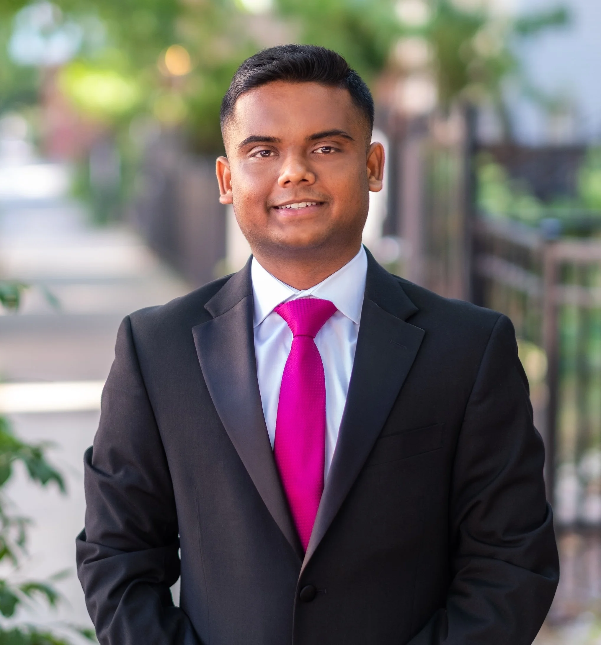 Professional Corporate Headshot by Minneapolis Photographer Jay Cupcake. Young man in a black suit and pink tie standing outdoors on a sunny day, with blurred trees and a sidewalk in the background.