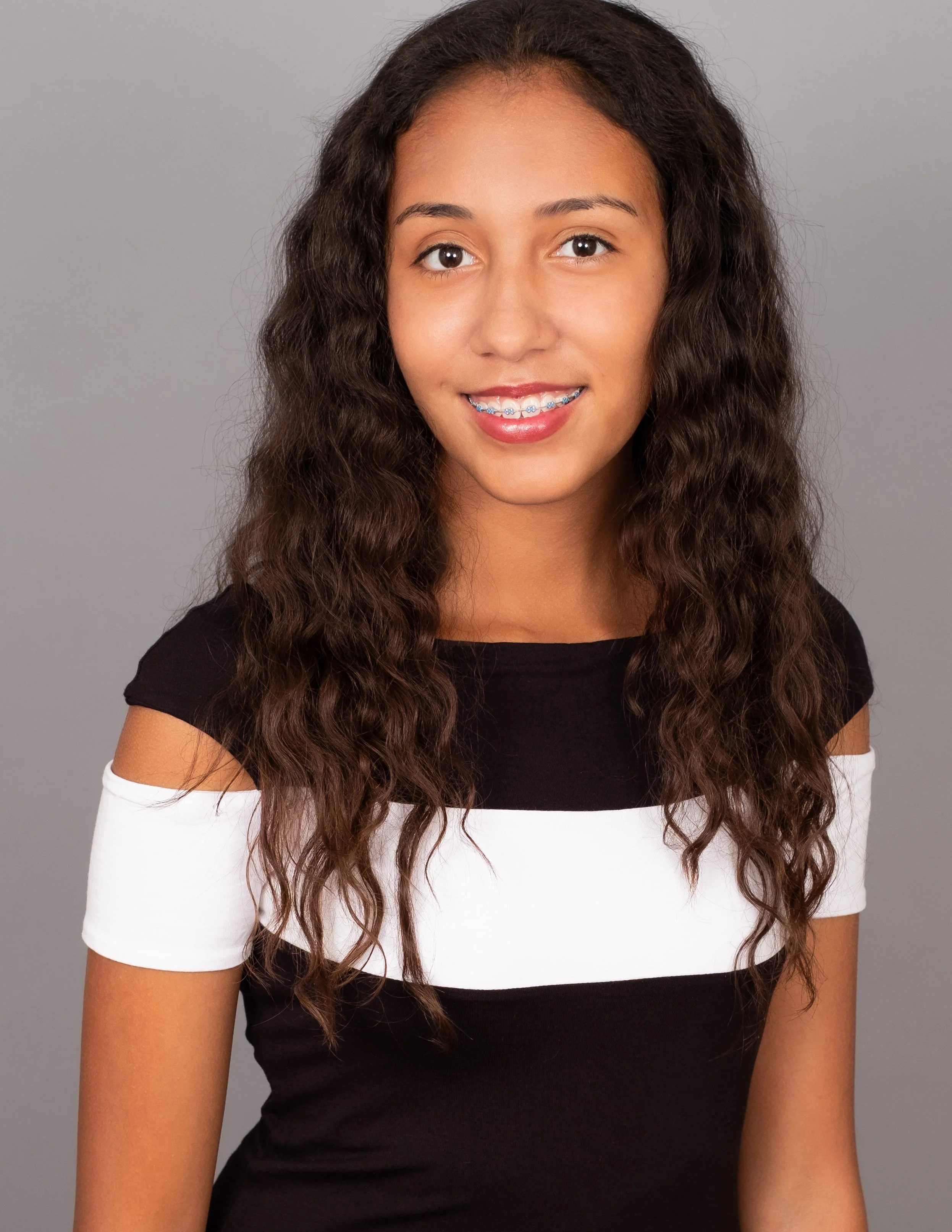Professional headshot by St. Paul photographer Jay Cupcake. Teenage girl with curly hair, braces, and a black and white off-shoulder top smiling against a gray background.