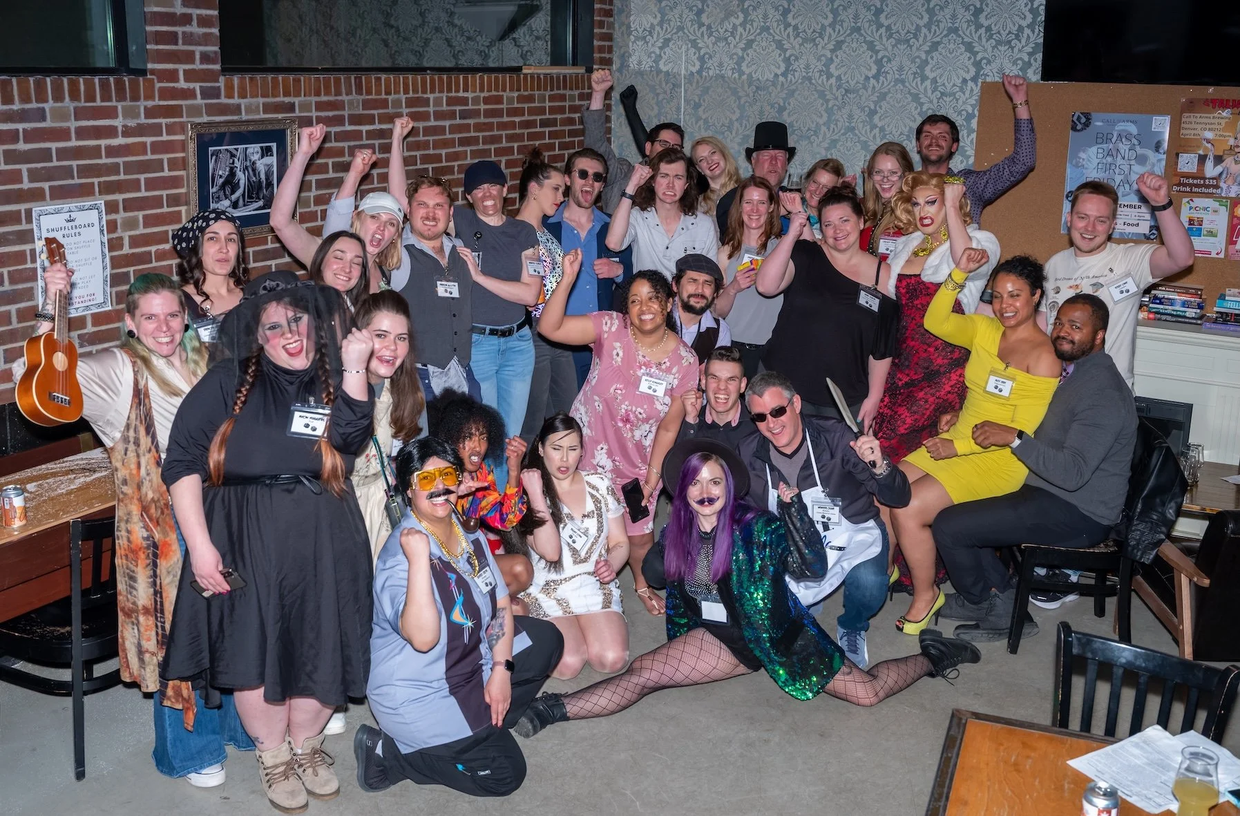 Professional Event Photography by Twin Cities Photographer Jay Cupcake. A large group of people dressed in colorful costumes and casual clothing, celebrating together in an indoor space with brick walls and patterned wallpaper. Some are wearing wigs,