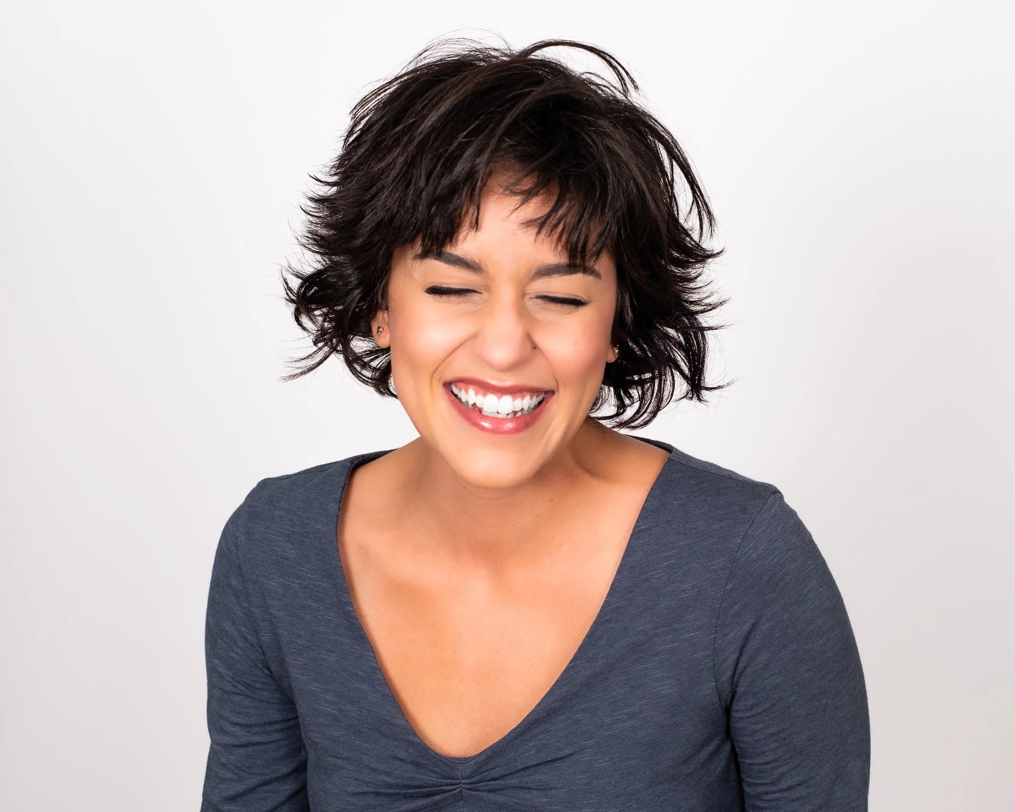 Professional Portrait Photography by Twin Cities Photographer Jay Cupcake. A woman with short, curly dark hair smiling and laughing with eyes closed, wearing a dark gray top, against a white background.