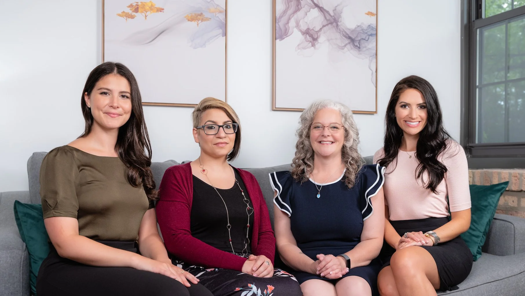 Professional Corporate Headshot by Twin Cities Photographer Jay Cupcake. Four women sitting on a gray couch in a living room, smiling at the camera. They have different hair colors and styles, and are dressed in casual and semi-formal clothing. Behin