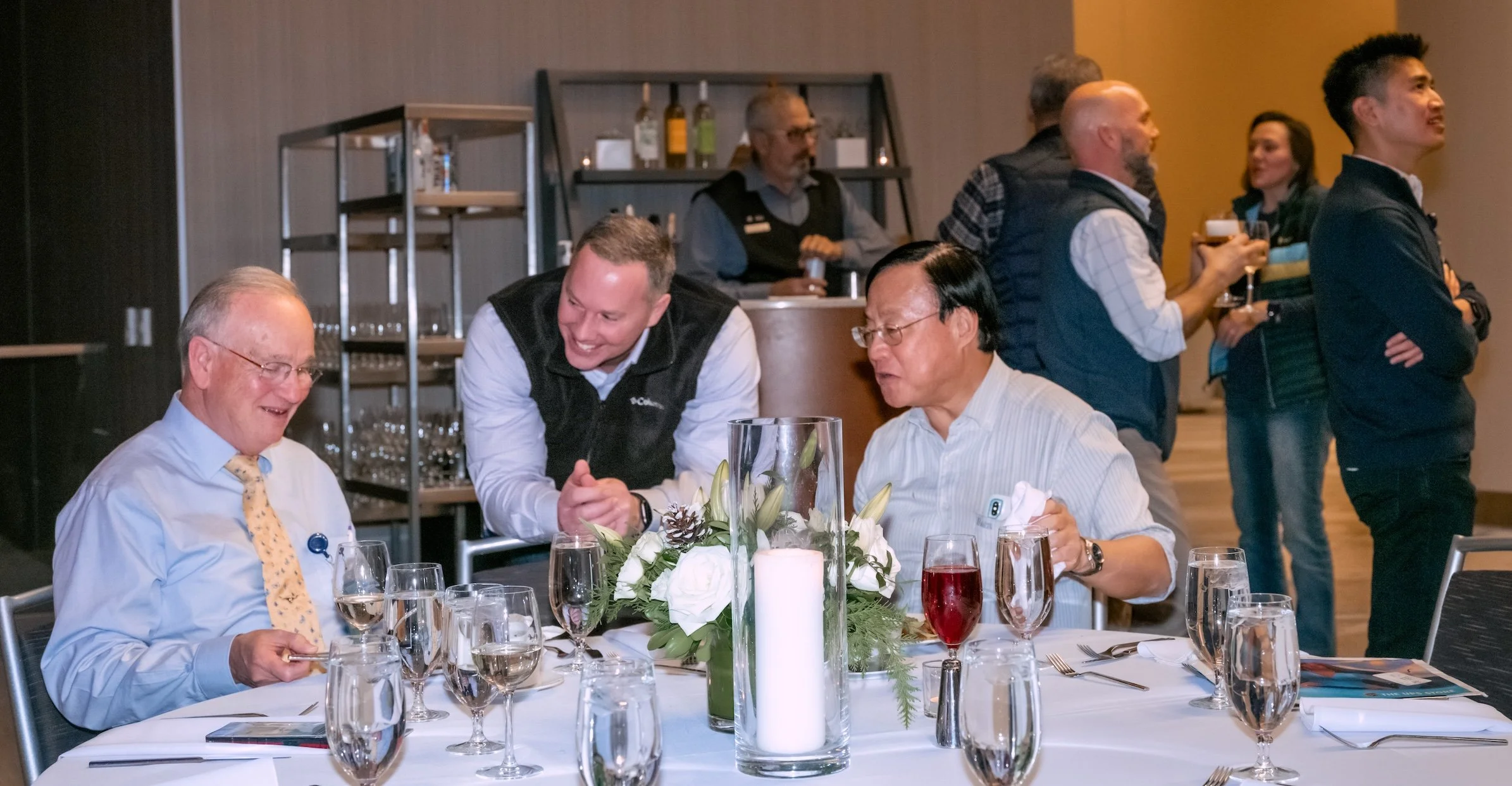 Professional Event Photography by Saint Paul Photographer Jay Cupcake. A group of men at a social gathering in a restaurant or banquet hall, sitting at a table with wine glasses and a floral centerpiece, engaging in conversation. In the background, m