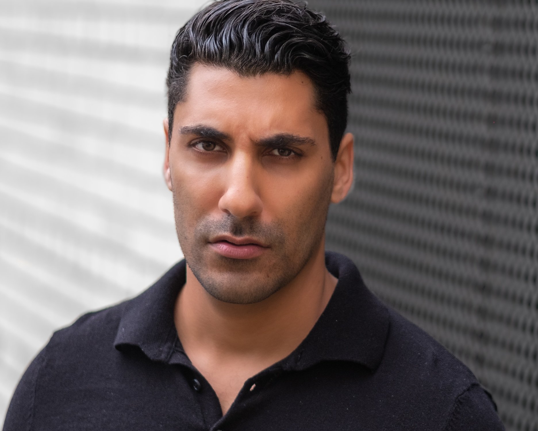 Professional Actor Headshot by Twin Cities Photographer Jay Cupcake. A man with dark hair and a serious expression, wearing a black shirt, standing outdoors near a textured wall.