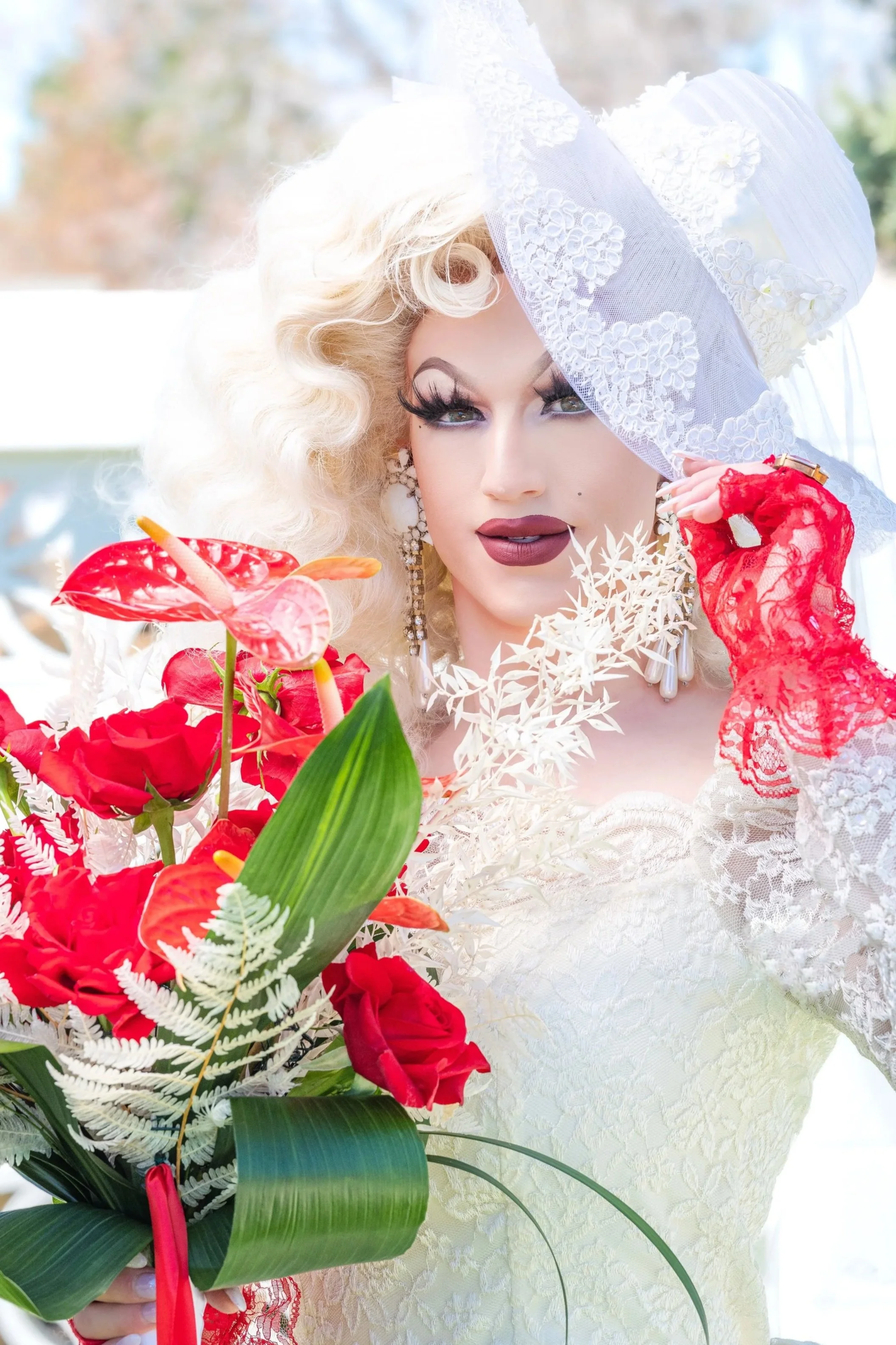 Professional headshot by St. Paul photographer Jay Cupcake. A person dressed in an elaborate white lace wedding gown and large hat, holding a bouquet of vibrant red flowers and wearing red lace gloves.