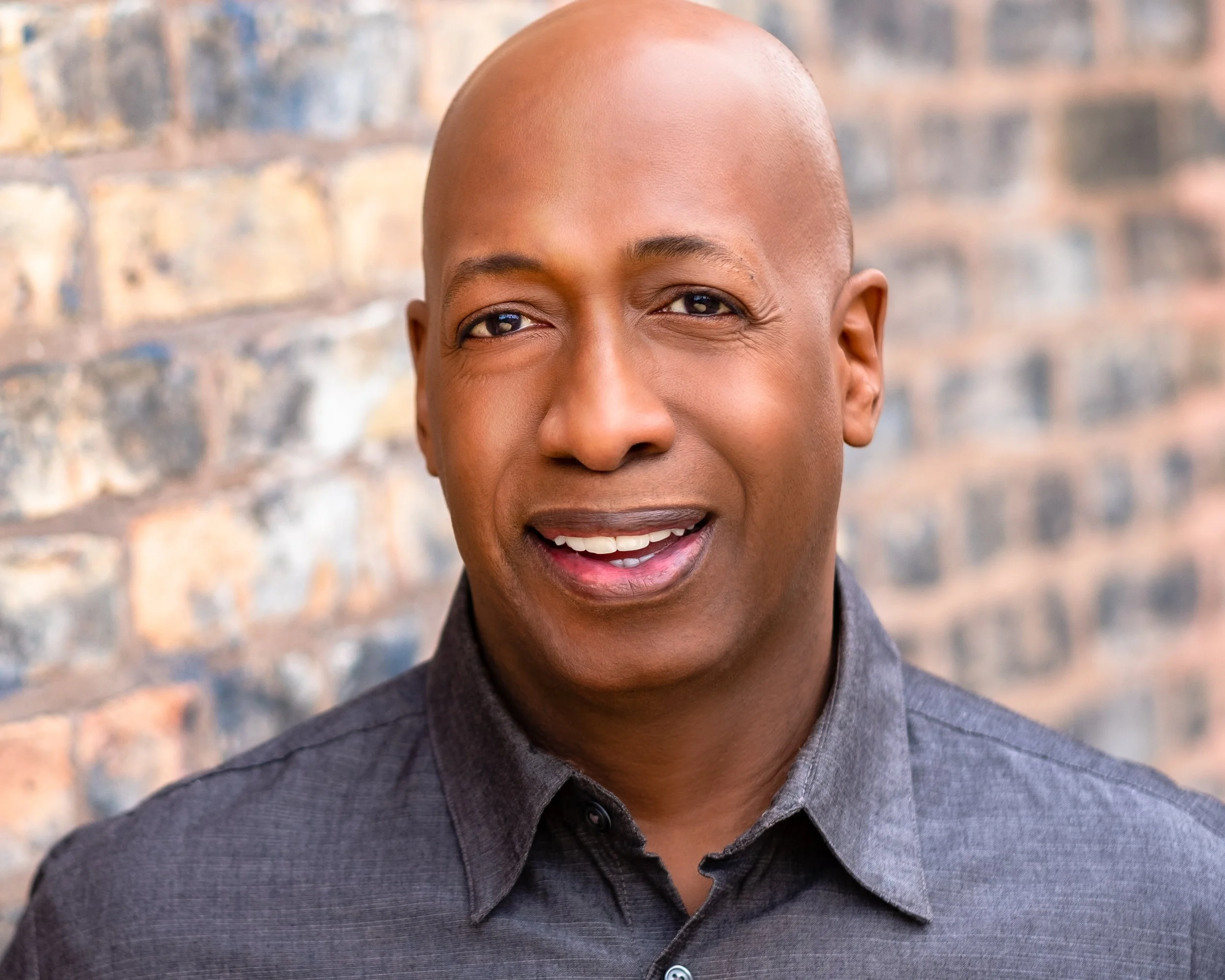 Professional headshot by St. Paul photographer Jay Cupcake. A smiling man with a bald head and dark eyes, wearing a dark gray button-up shirt, standing in front of a brick wall.