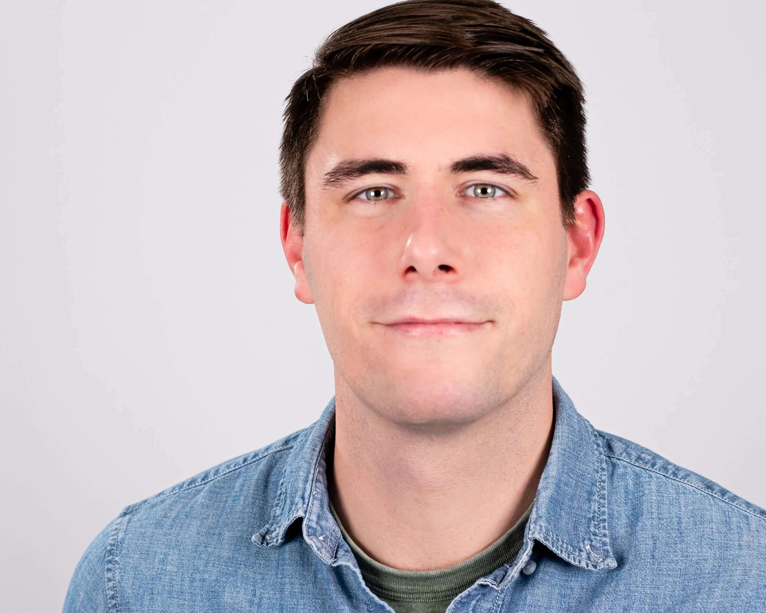 Professional headshot by St. Paul photographer Jay Cupcake. A close-up portrait of a young man with dark brown hair, light-colored eyes, wearing a denim shirt, against a plain light gray background.